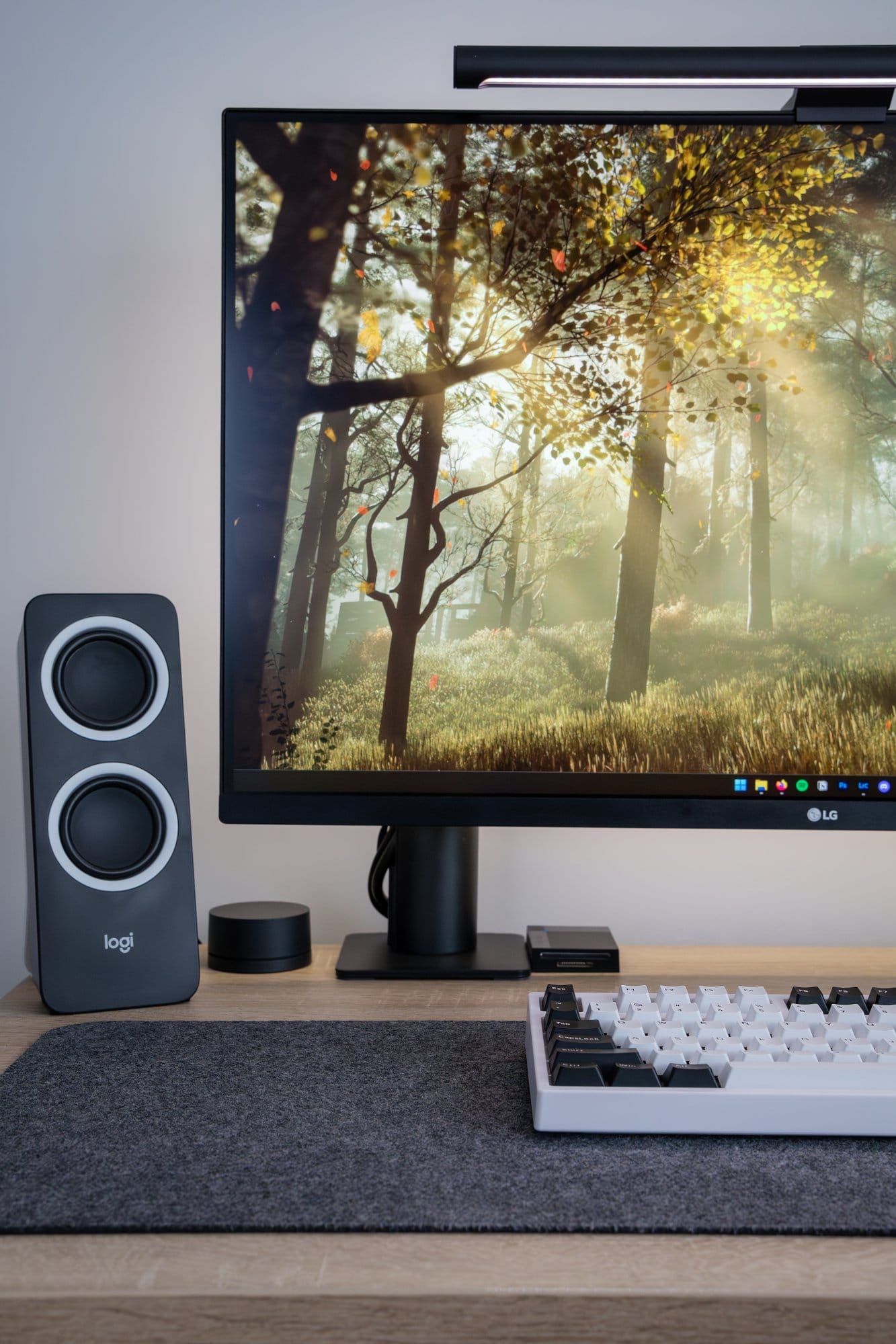 A close-up of the minimal workspace featuring an LG monitor with a forest background, a black stereo speaker, a minimalist soundbar, and a mechanical keyboard on a felt mat