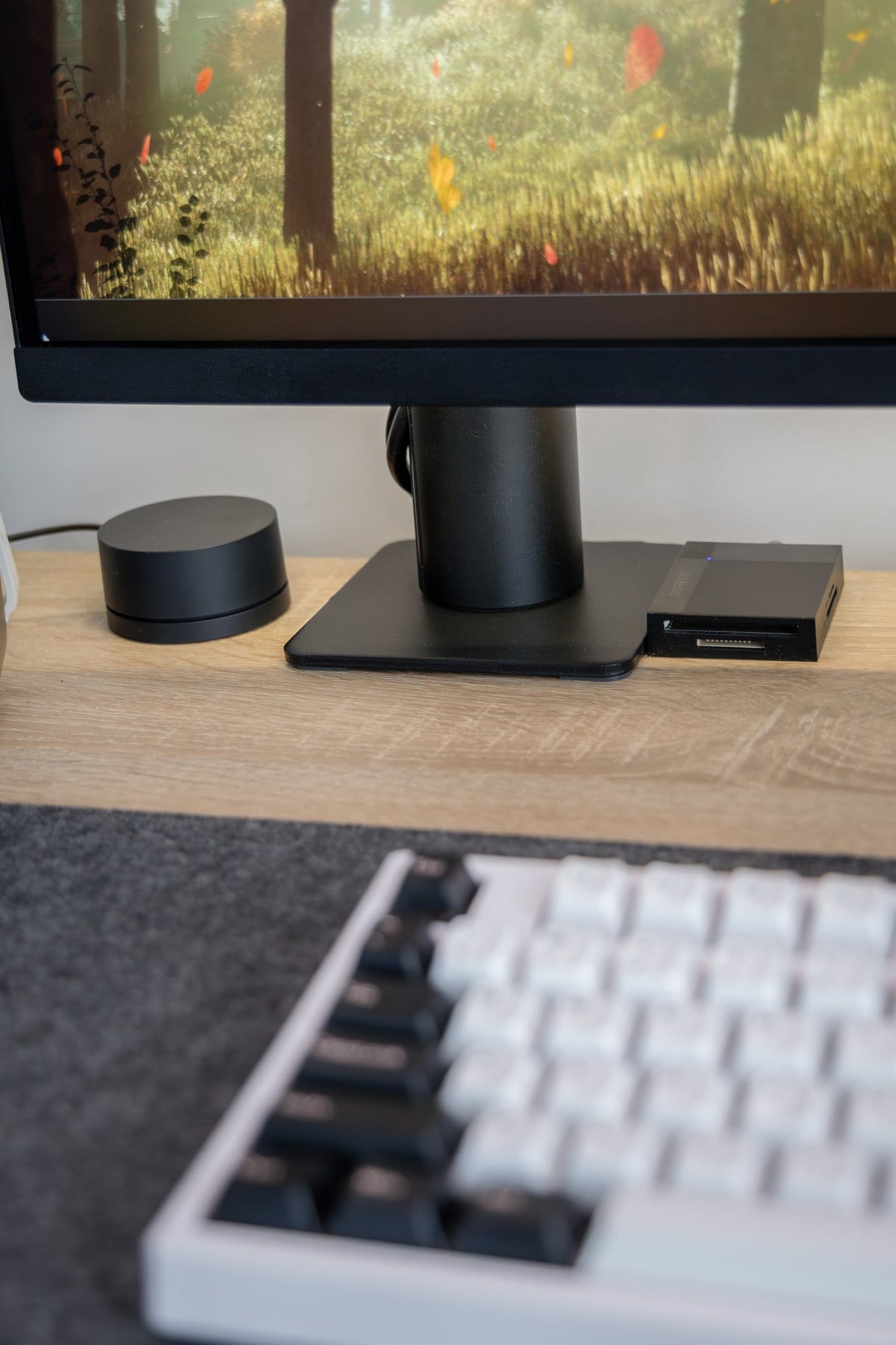 A desk setup detail showing the base of a monitor, a soundbar, and a sleek external drive, with the corner of a white mechanical keyboard in the foreground, all atop a wooden desk with a grey desk mat