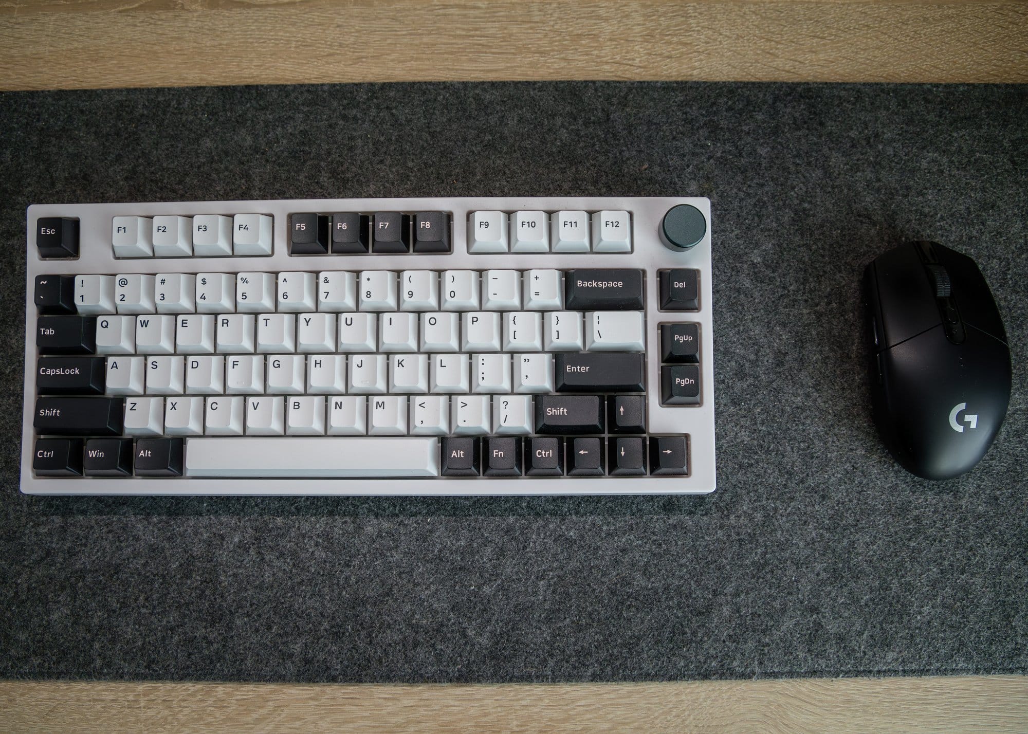 Top-down view of a mechanical keyboard with black and white keys alongside a wireless black gaming mouse, both resting on a grey felt desk mat on a wooden surface