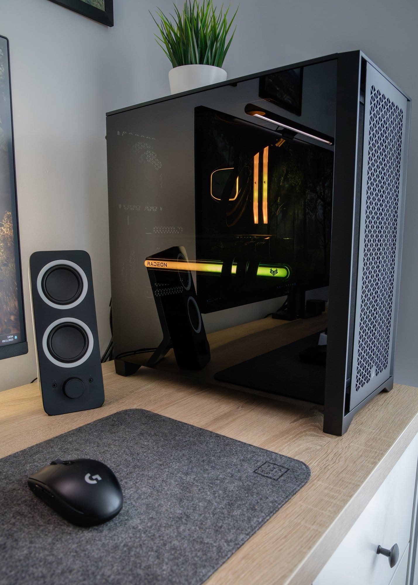 A close-up of a high-end custom-built gaming PC with a transparent side panel revealing internal components, illuminated by LED lights, placed on a desk beside a black stereo speaker, with a wireless mouse on a grey mat and a potted plant on top of the case