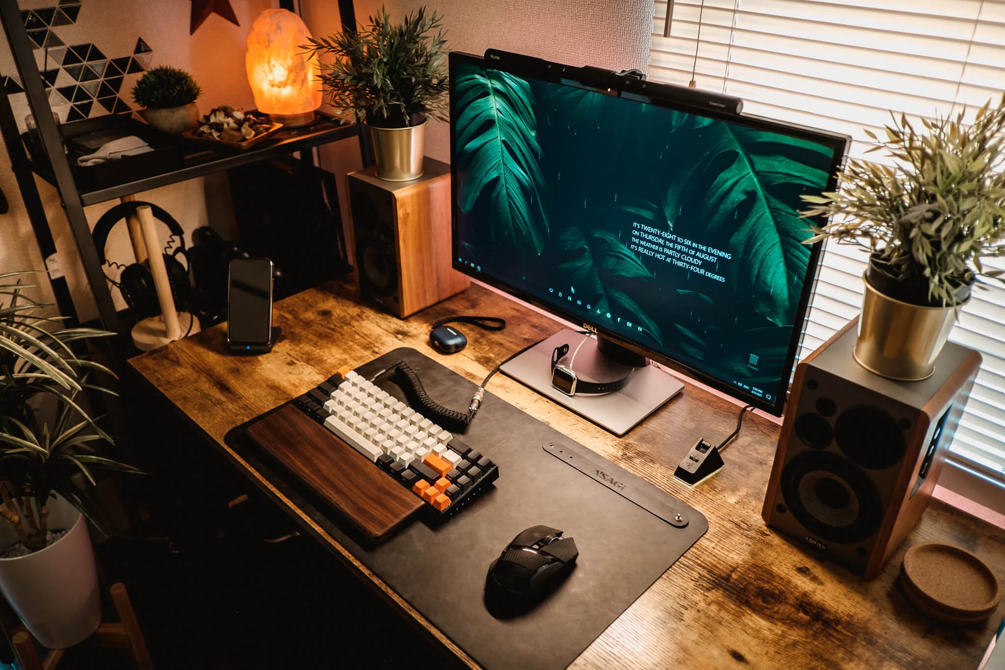  A well-organised desk setup with a glowing salt lamp, multiple plants, a mechanical keyboard, a large monitor, and a desk mat, set in a room with warm, ambient lighting