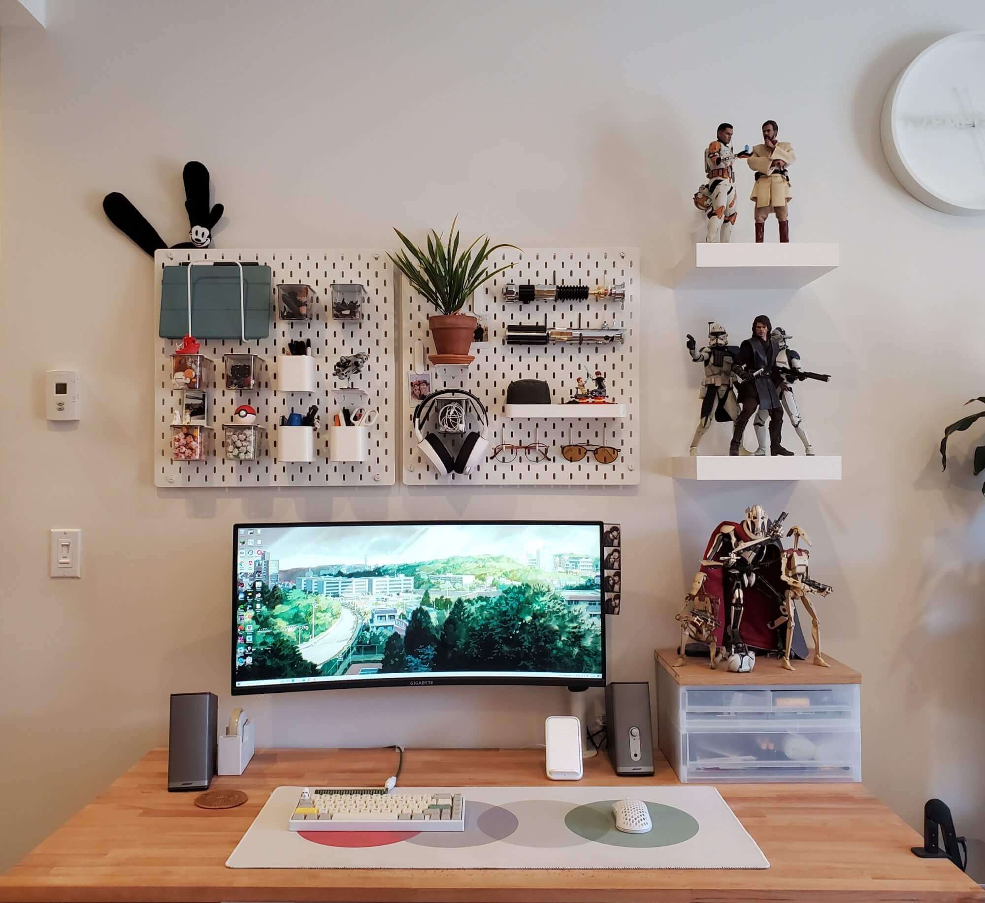 A well-organised desk with an ultrawide monitor, flanked by pegboards and decorative shelves displaying collectible figures, plants, and miscellaneous items, in a room with a minimalist aesthetic