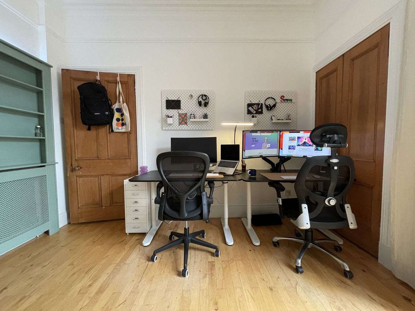 A shared home office featuring two standing desks with ergonomic chairs and computers, a backpack and a tote bag hanging on the door, and pegboards with various items on the wall