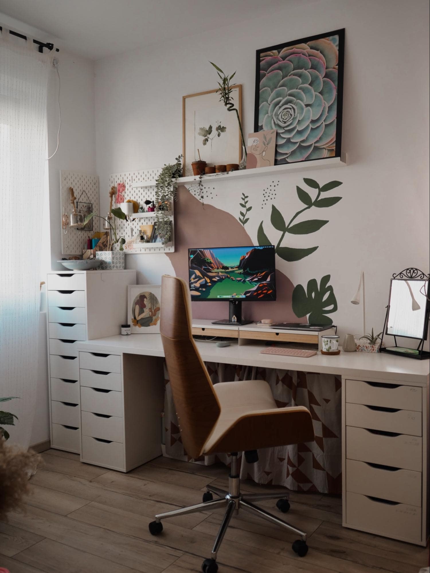 A cosy, artistic home office with a computer on a white desk, surrounded by plants and pegboards, an ergonomic chair, IKEA ALEX drawers, and a large succulent painting on the wall