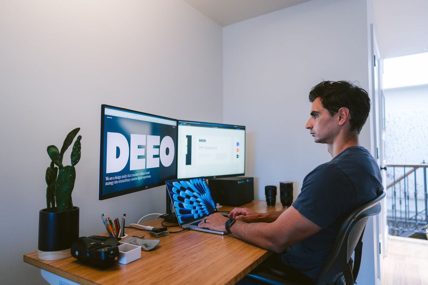 A person is seated at a desk working on a laptop, flanked by two large monitors displaying bold graphics, with office accessories and a potted plant nearby