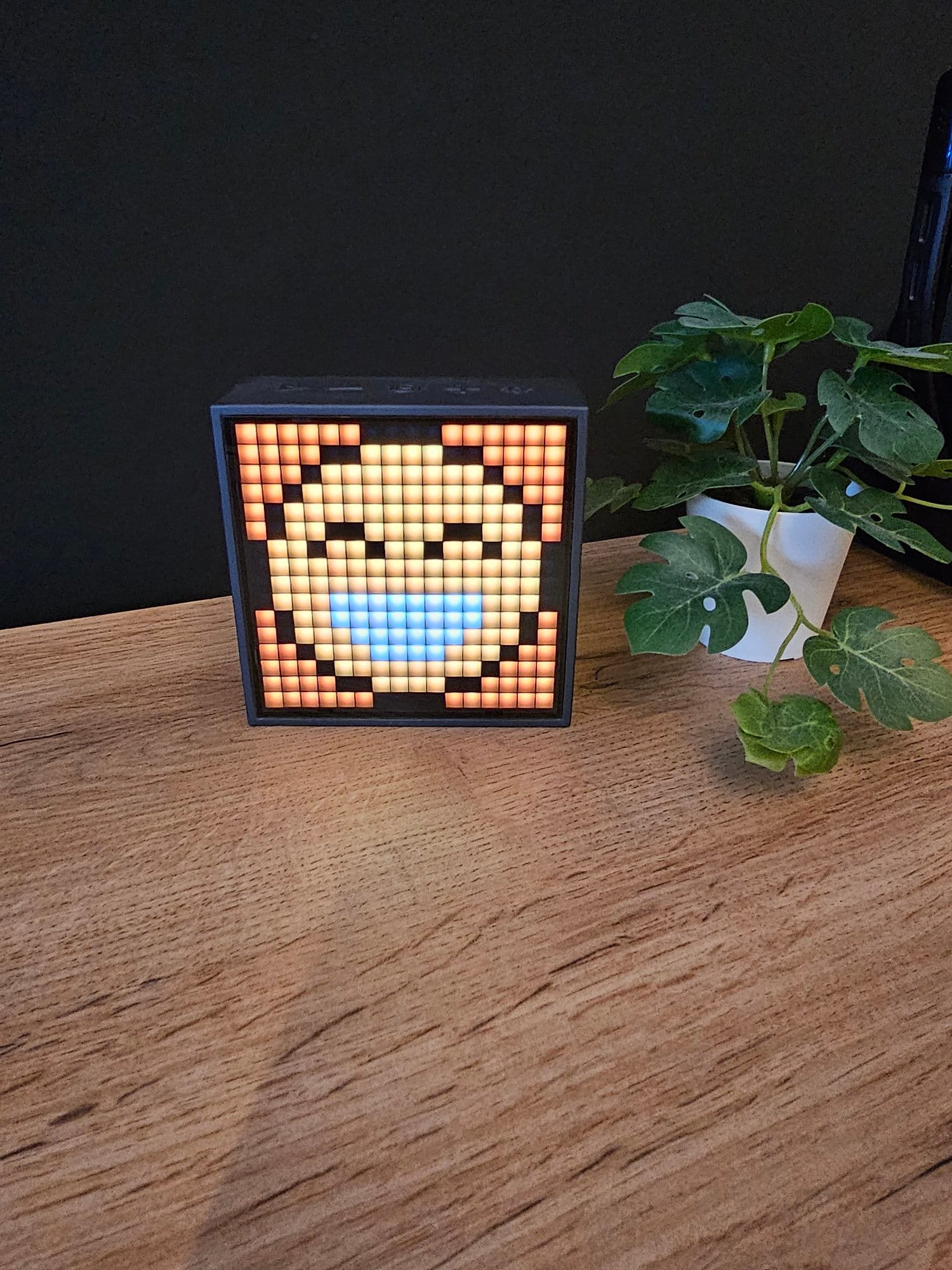 A Divoom TimeBox Evo speaker next to a potted green plant on a wooden surface against a dark wall