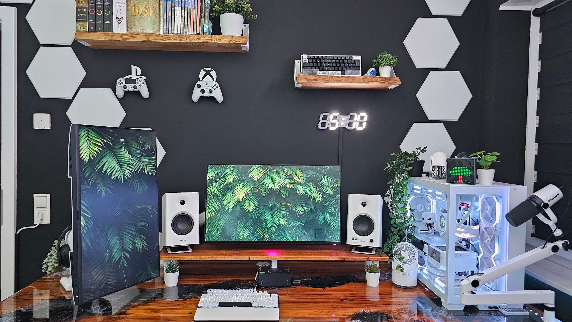 A well-organised gaming station featuring a decorative black wall with mounted hexagon shapes, gaming controllers, a digital clock, and floating shelves with books and plants, complemented by a vibrant dual-monitor setup and a white-themed custom PC
