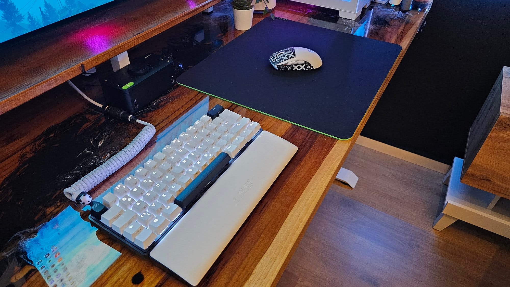 A close-up view of a modern workspace featuring a white mechanical keyboard on a large mouse pad, a custom-designed gaming mouse, and various tech gadgets on a polished wooden desk with ambient lighting in the background