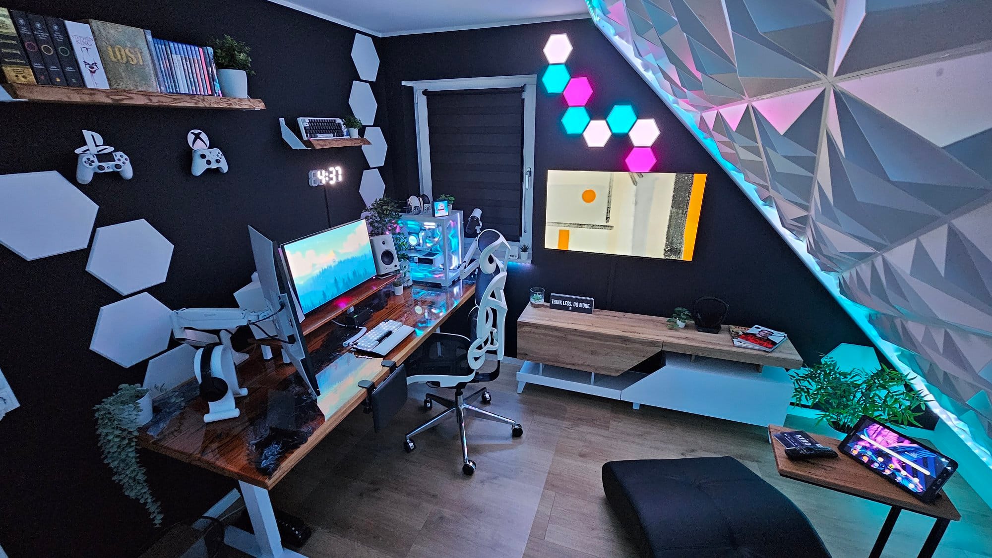 A panoramic view of a dark-themed gaming room with ambient lighting, featuring a multi-monitor PC setup, decorative acoustic panels, floating shelves with books, gaming controllers on the wall, and a cosy lounge chair