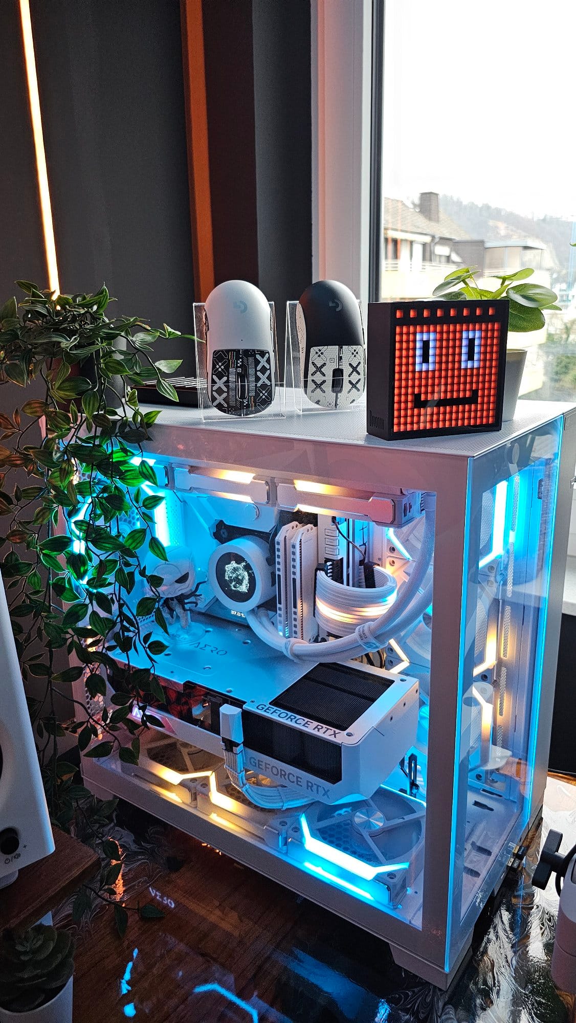 A close-up of a custom gaming PC with blue LED lighting, showcasing high-end components and liquid cooling, accompanied by decorative figures and a pixel art display, set beside a window with a view