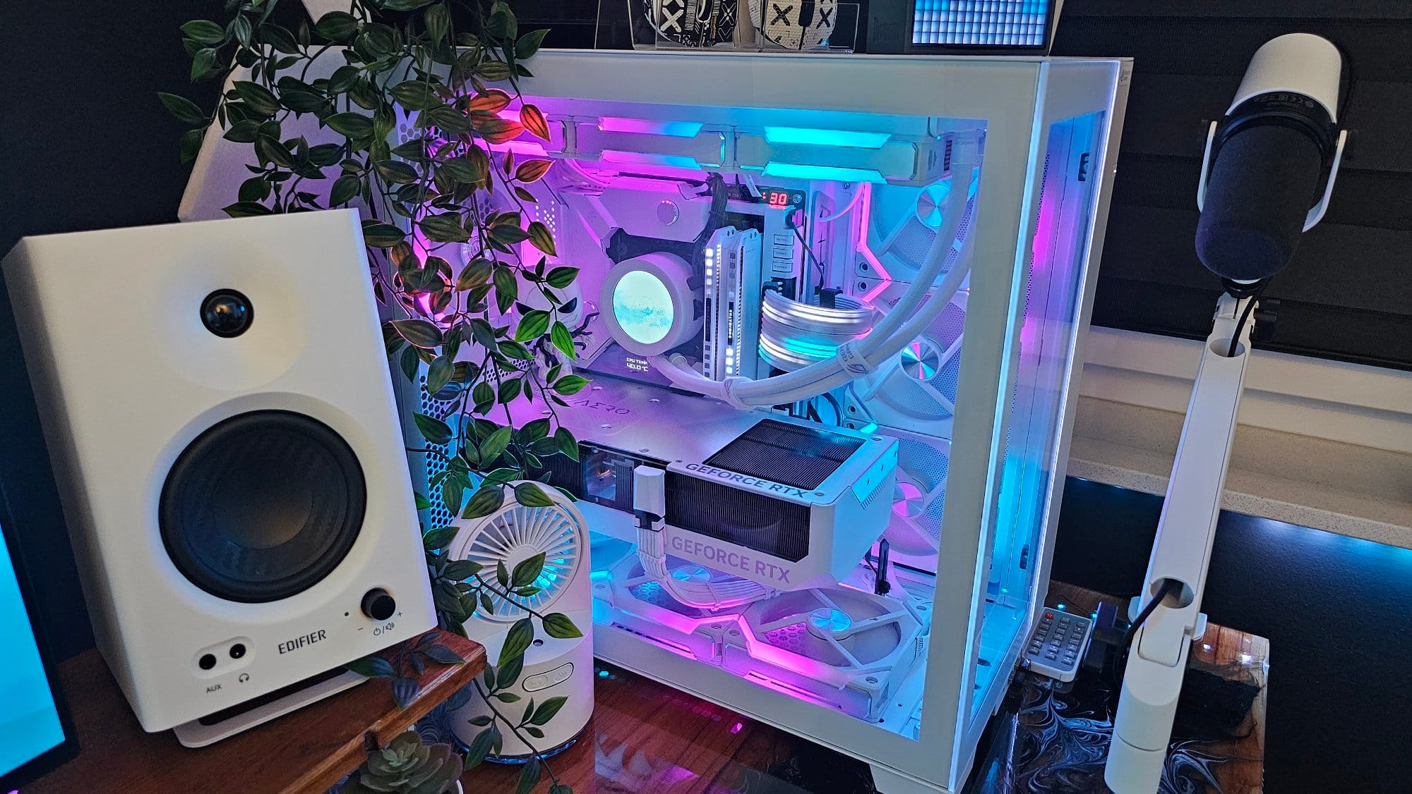 A close-up of a high-end custom PC with RGB lighting, situated next to a speaker and a microphone on a boom arm, with a potted plant adding a touch of greenery to the modern tech setup