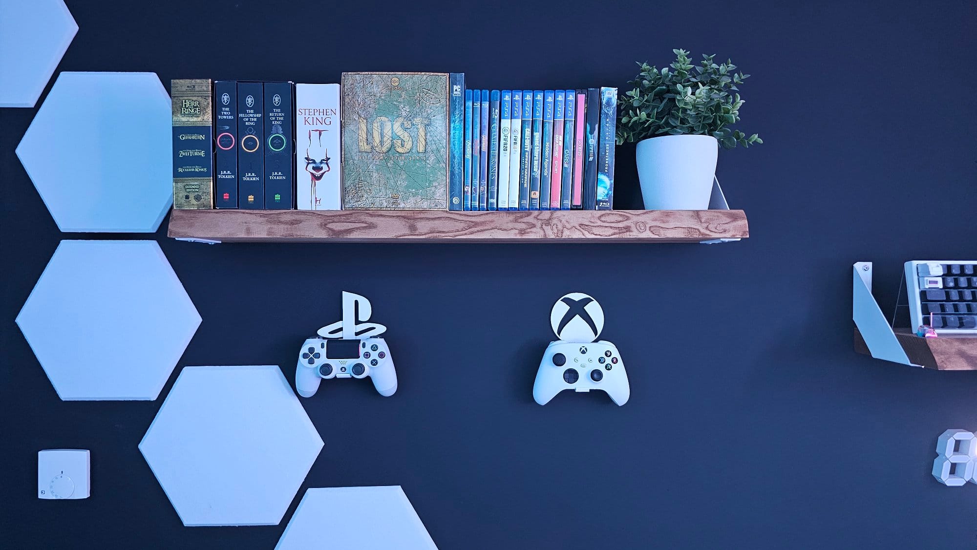 A dark-coloured wall decorated with a wooden shelf holding books, Blu-ray cases, a potted plant, and mounted hexagonal shapes, along with gaming controllers representing PlayStation and Xbox