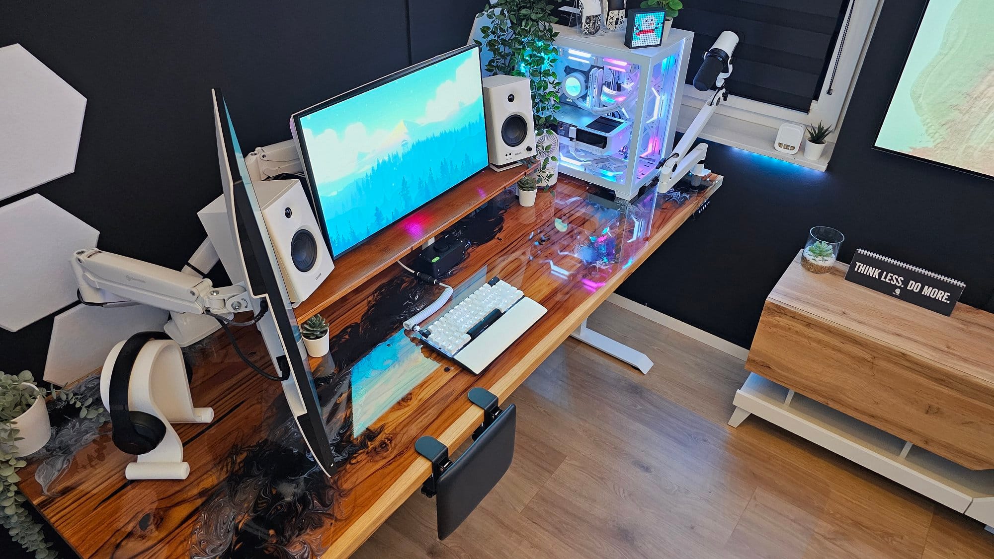 An elevated view of a gaming and work setup with a vibrant dual-monitor display, speaker system, and a high-end computer with RGB lighting, all on a polished wooden desk accented by modern accessories and a desk sign that reads “THINK LESS. DO MORE”