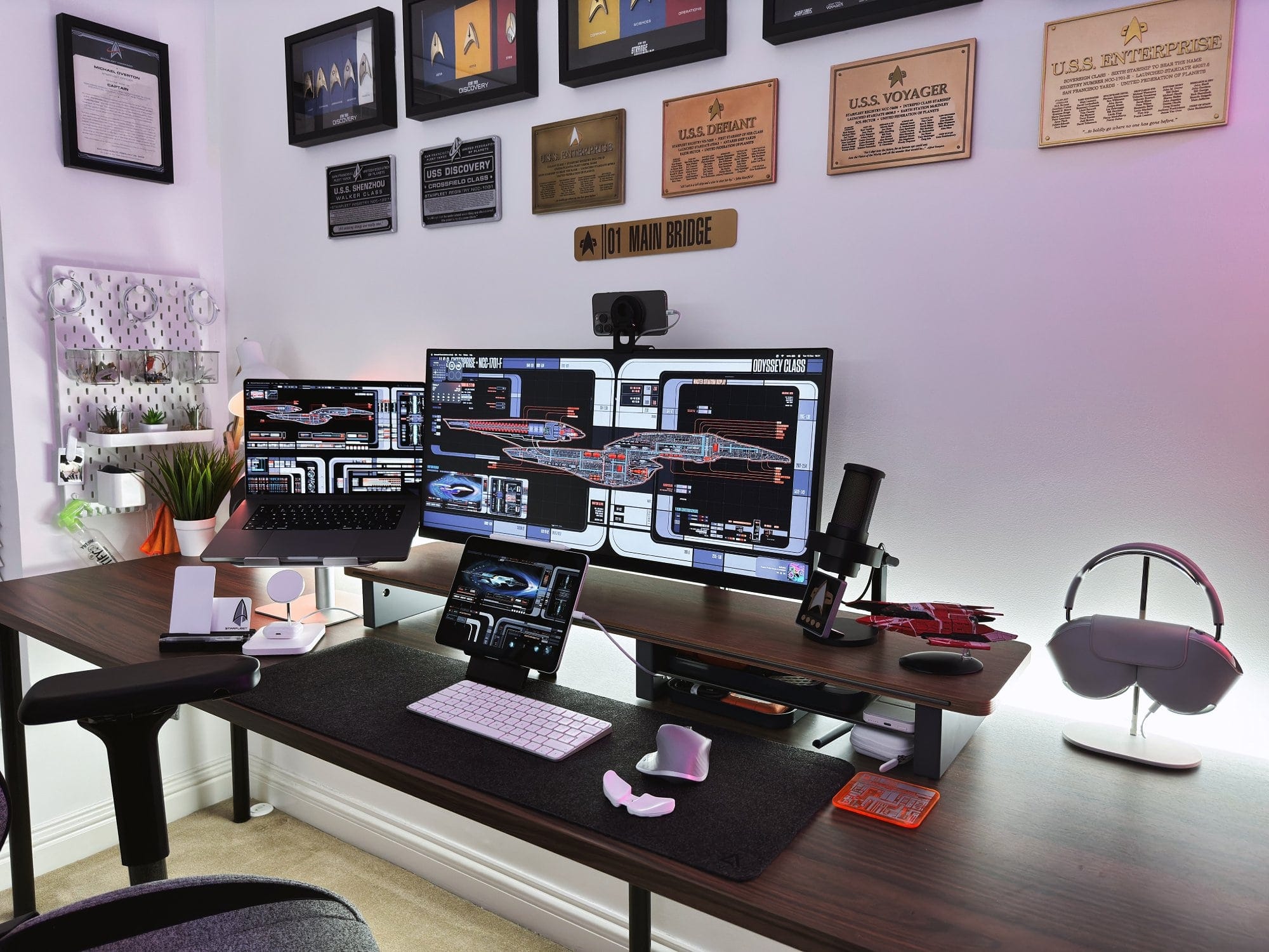 A sleek workspace with a multi-monitor setup featuring Star Trek graphics, a laptop, tablet, microphone, and headphones, with Star Trek memorabilia in the background