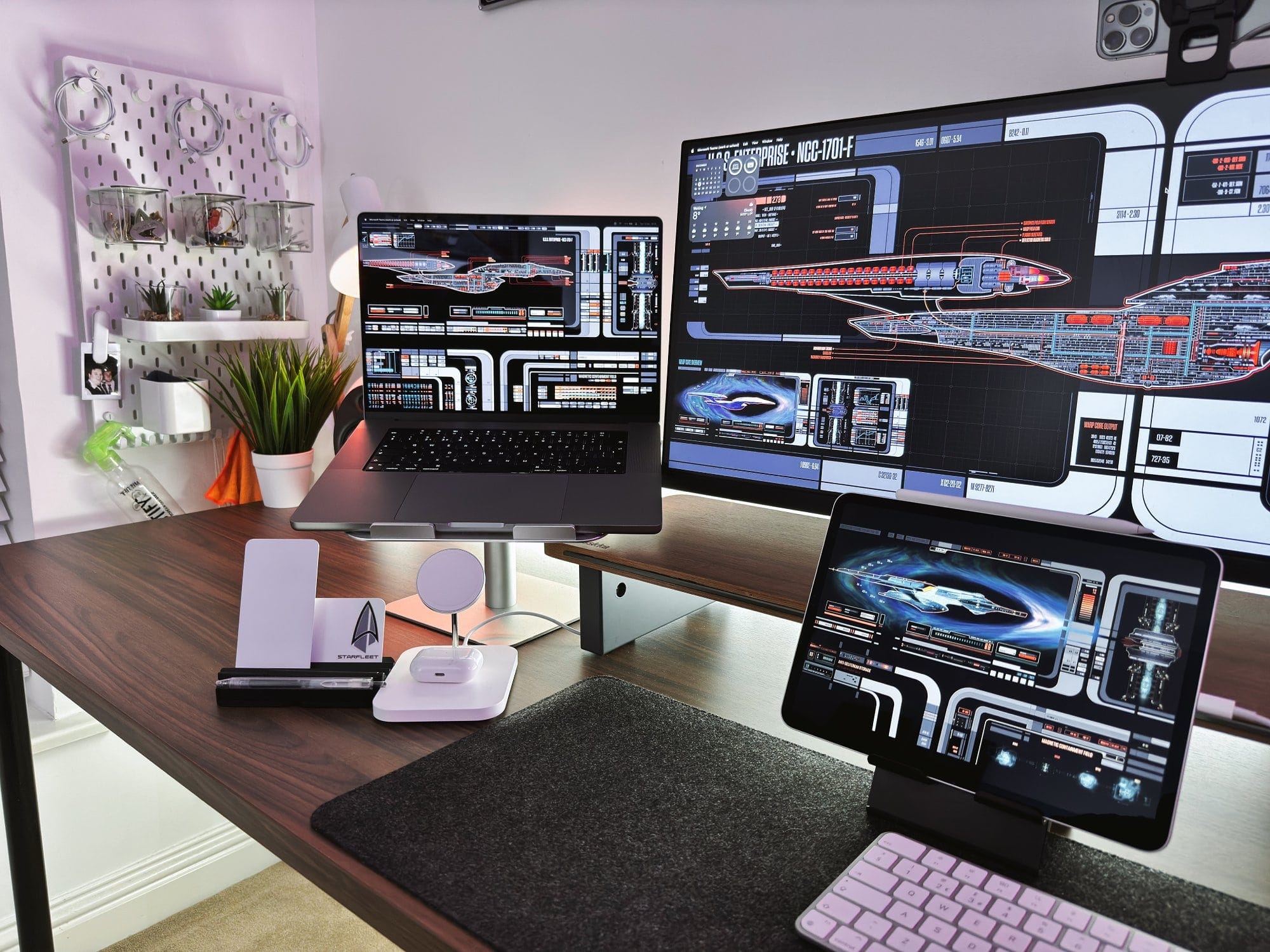 A modern desk setup with a laptop and tablet displaying Star Trek starship schematics, accompanied by a pegboard on the wall