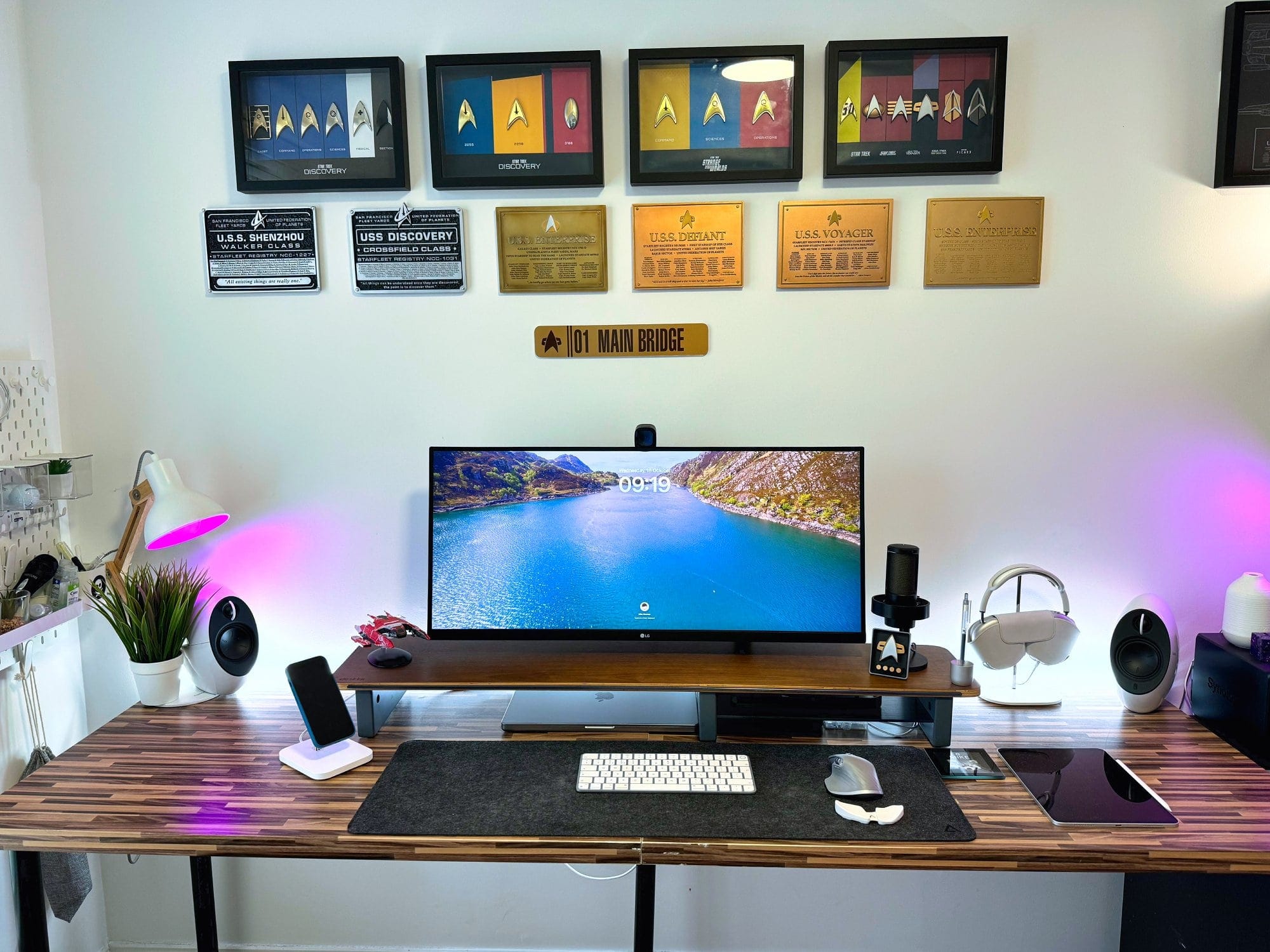 A well-lit home office setup with a wooden desk, a single monitor, ambient lighting, and a wall adorned with framed Star Trek insignia and plaques