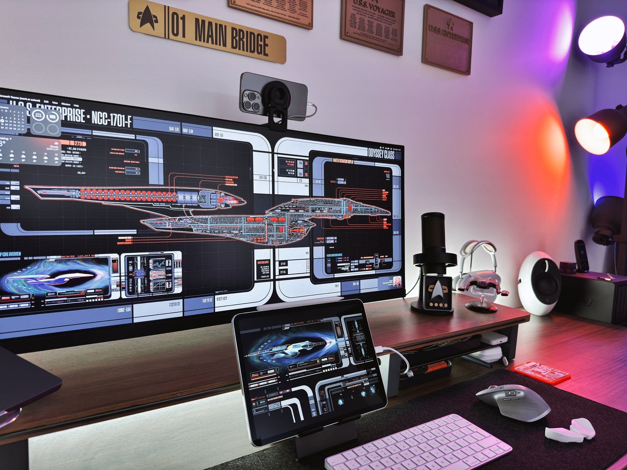 A high-tech desk setup with a multi-monitor display showing Star Trek interfaces, a tablet on a stand, ambient coloured lighting, and Star Trek memorabilia on the wall