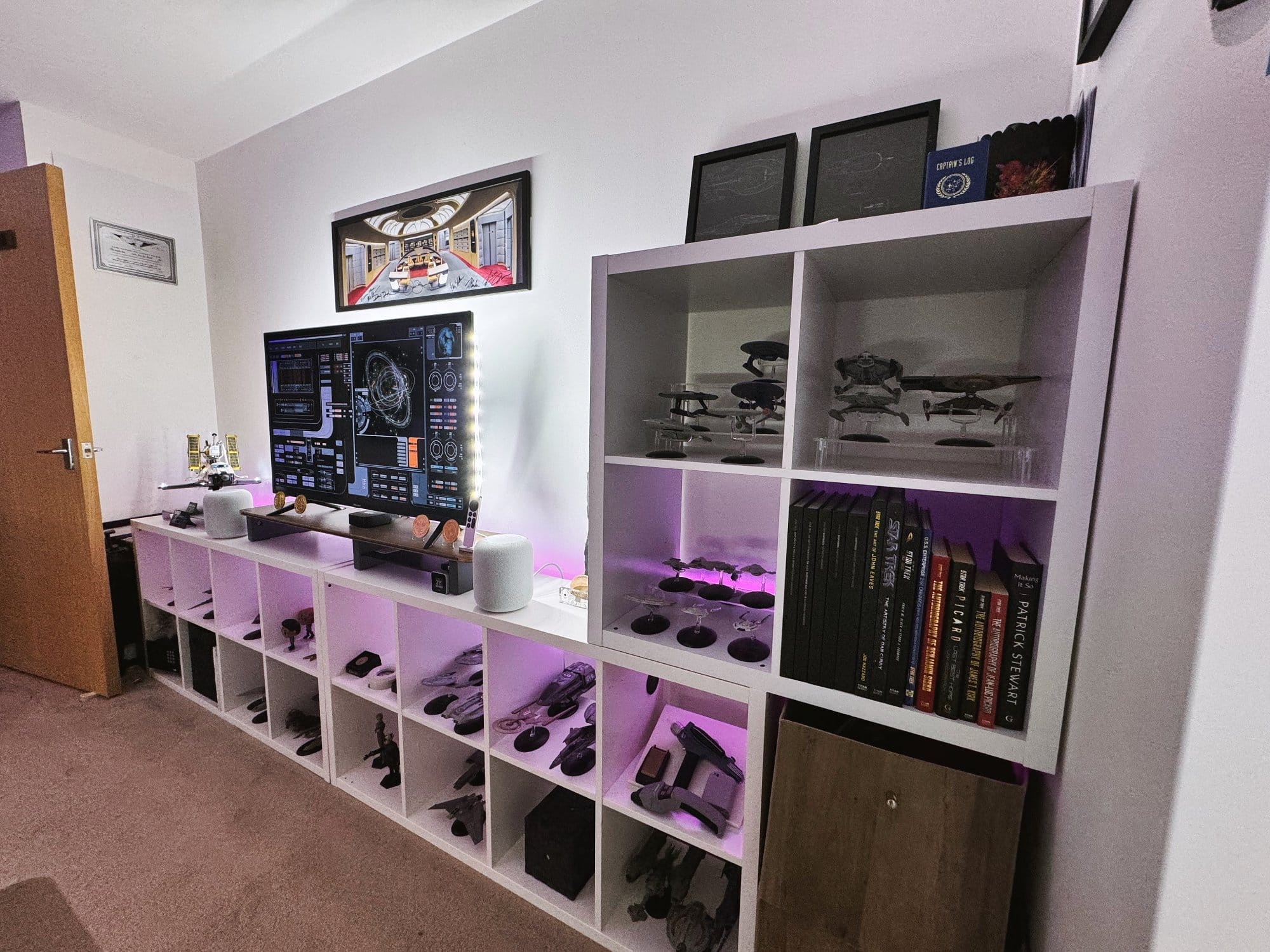 A home office with a large monitor, ambient lighting, and a shelving unit displaying a collection of Star Trek models and books, with framed artwork and certificates on the wall