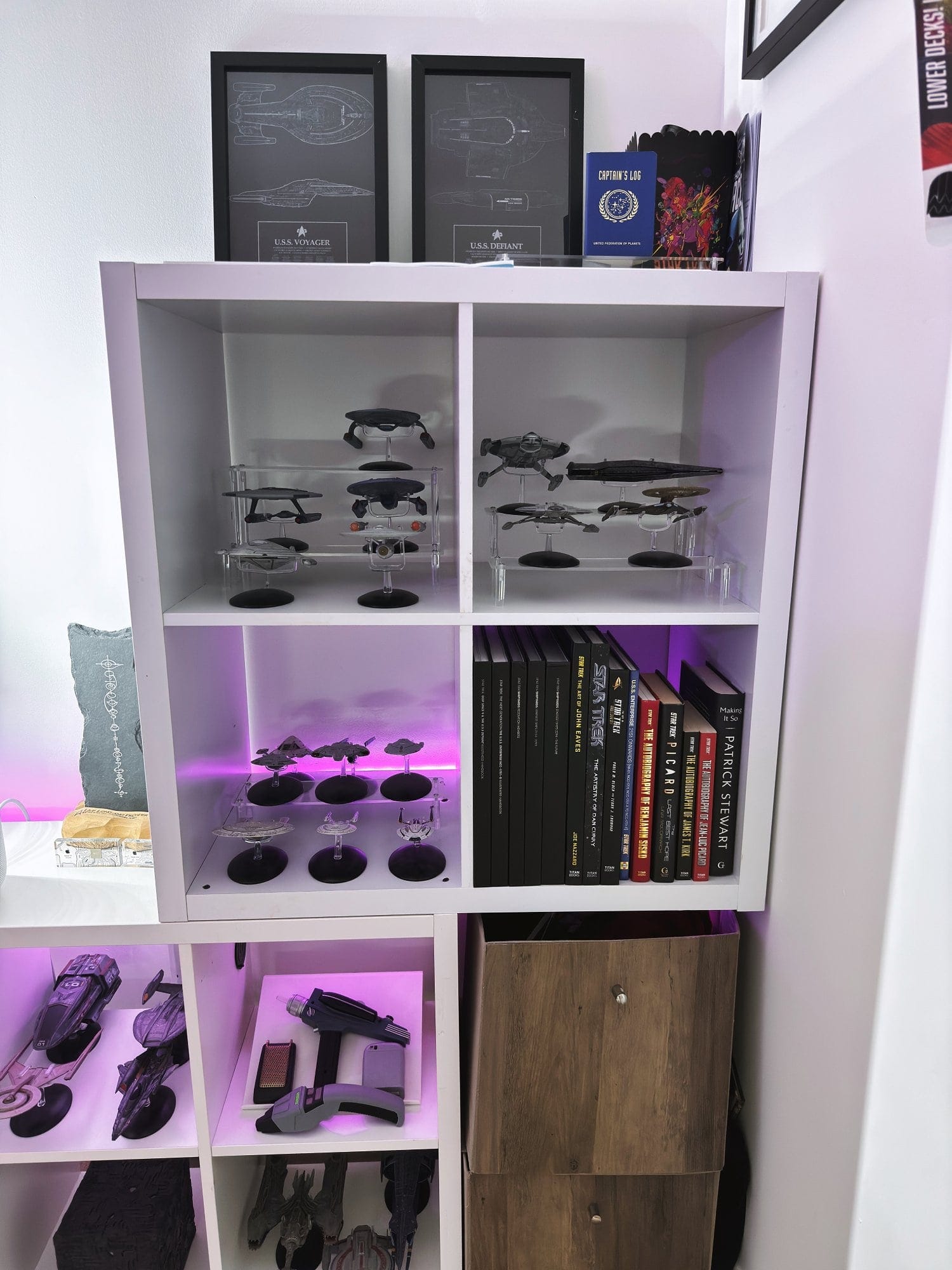 A vertical bookshelf displaying Star Trek spacecraft models on acrylic stands, flanked by framed blueprints and a collection of hardcover books