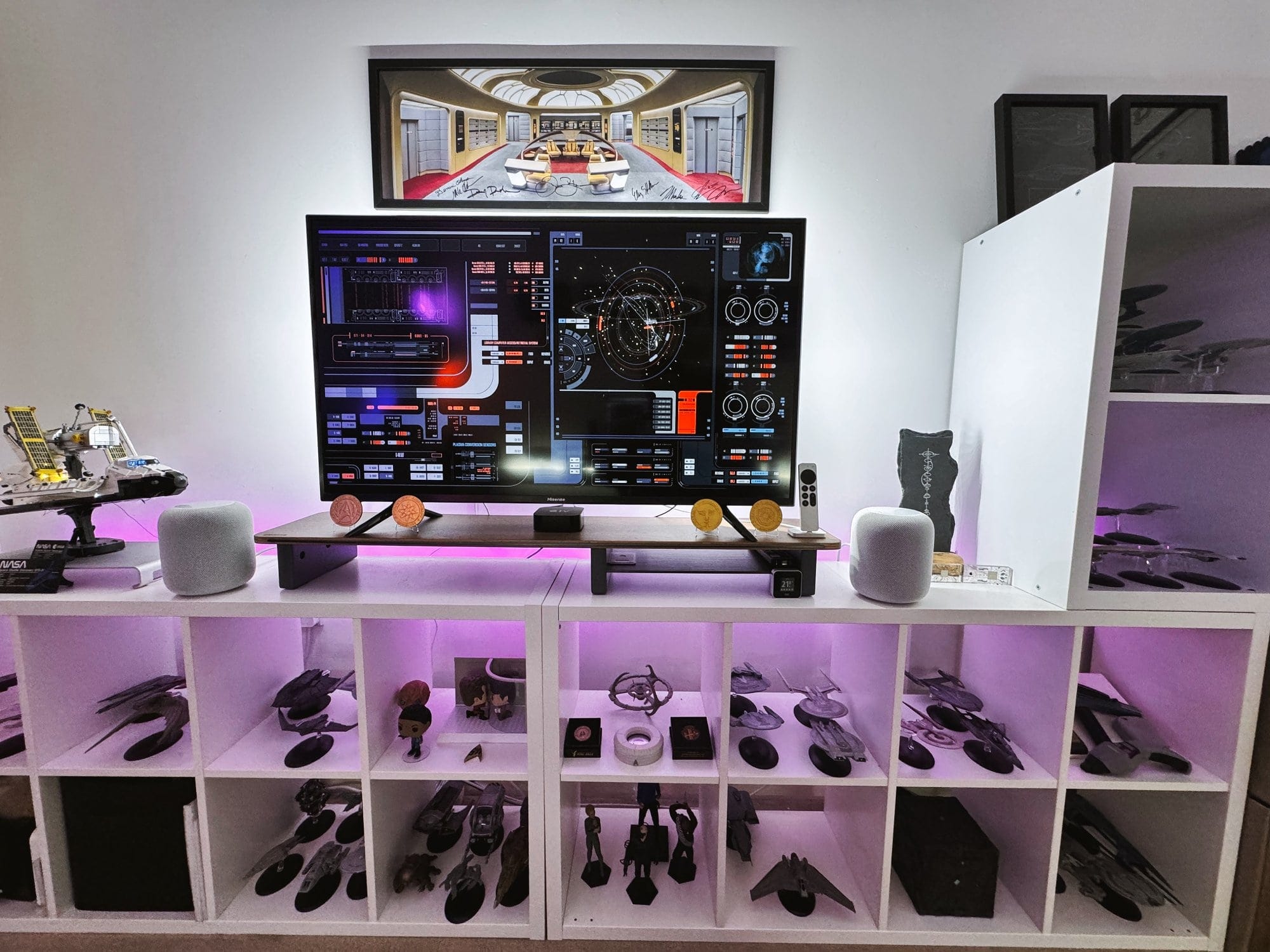 A close-up of a home office setup with a large display screen, various Star Trek starship models on a white shelving unit, and sci-fi themed artwork above the monitor