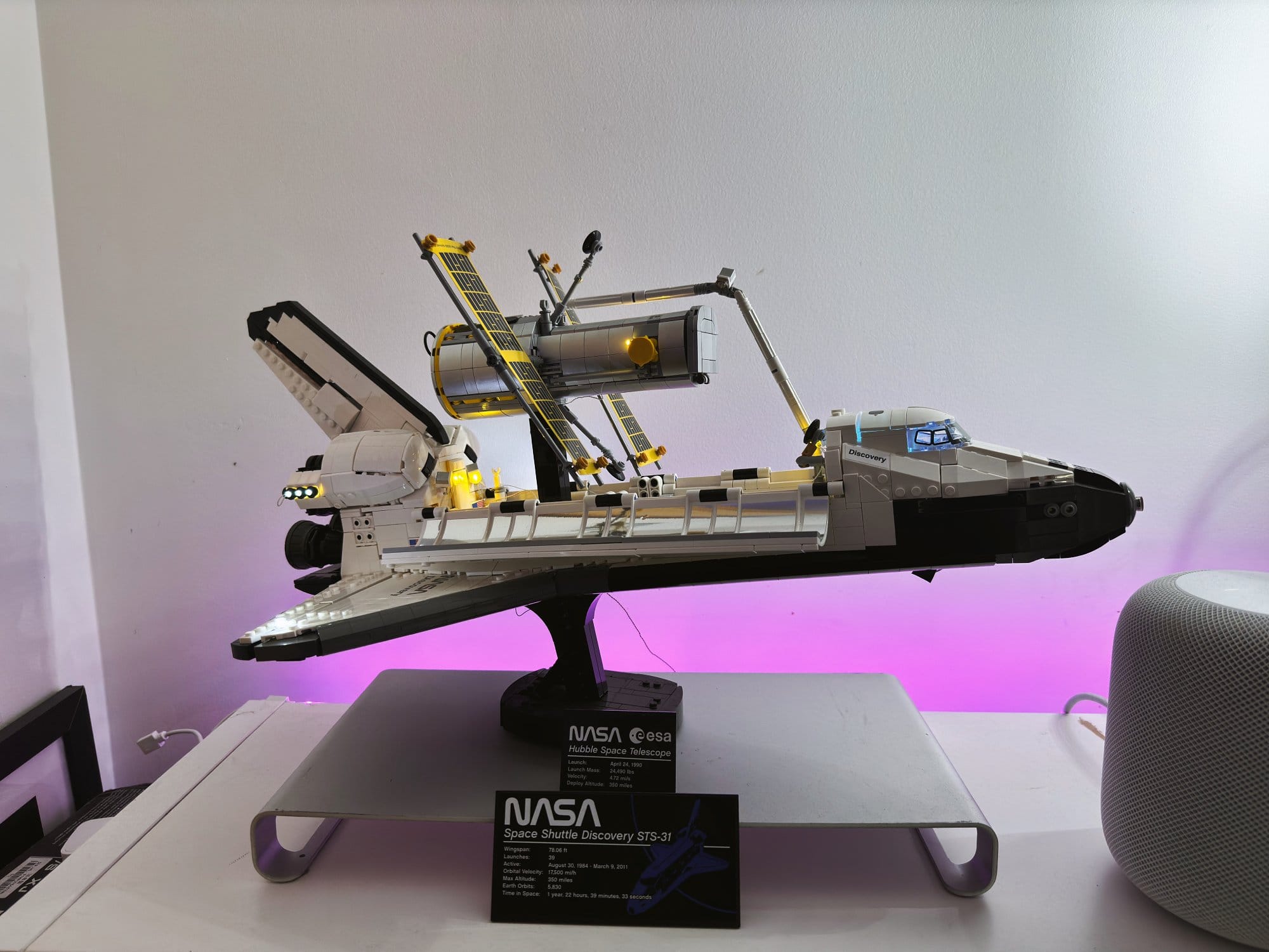 A detailed LEGO model of the NASA Space Shuttle Discovery on a stand, displayed against a backdrop of ambient purple lighting