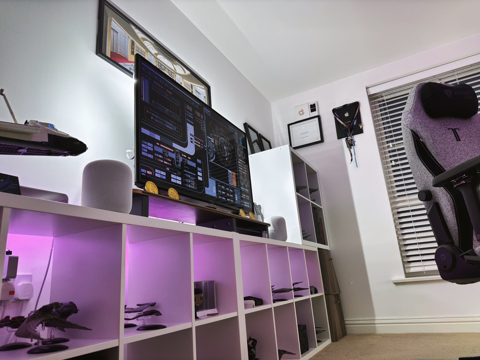 A corner view of a home office with a monitor on a white shelf displaying starship schematics, a gaming chair to the side, Star Trek models on the shelves, and certificates on the wall, all under a soft purple glow