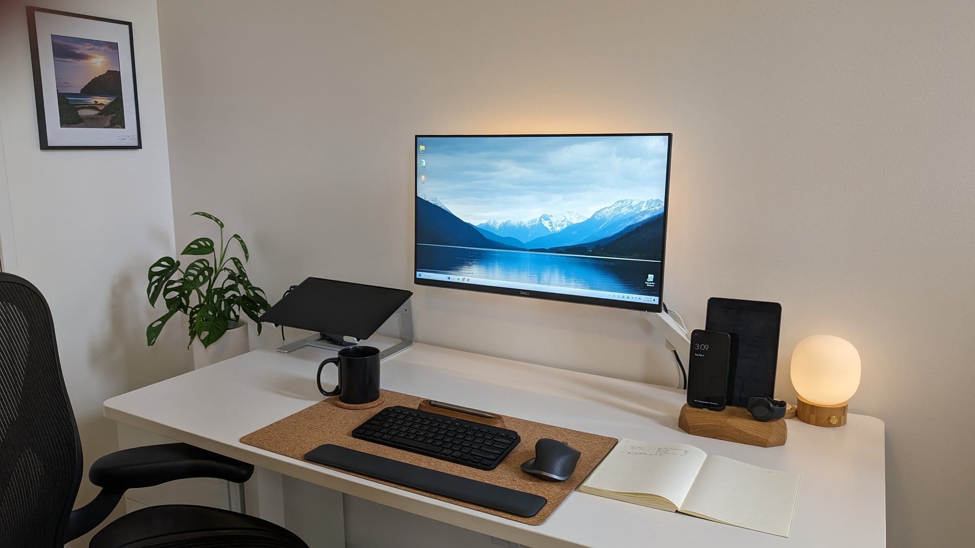 A home office setup with a monstera plant, a glowing orb lamp, and a framed landscape picture on the wall