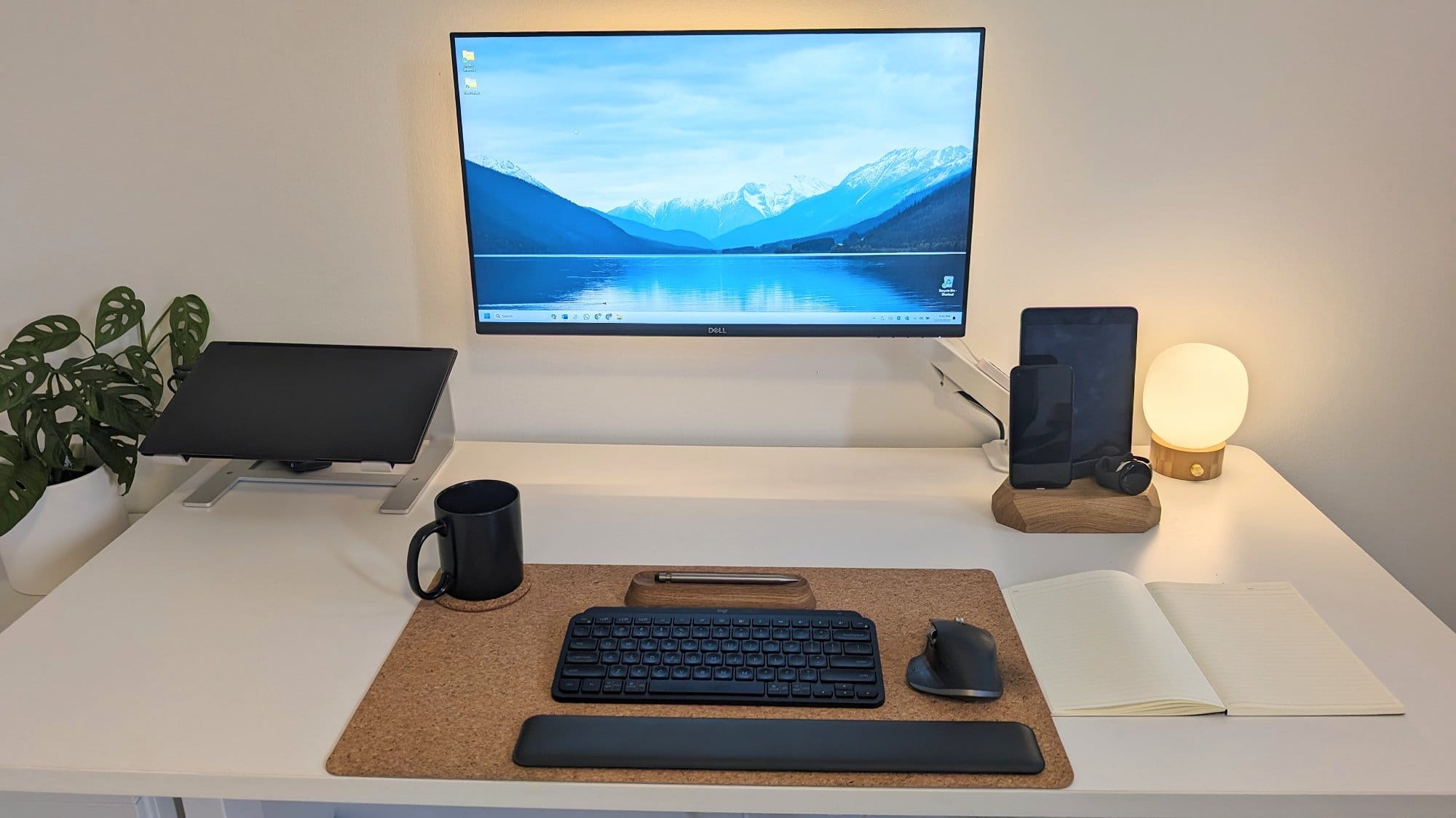 A well-arranged workstation with a large monitor, a laptop on a stand, a black mug, a wireless keyboard and mouse on a cork mat, as well as a tablet and smartphone on a wooden Oakywood charging dock