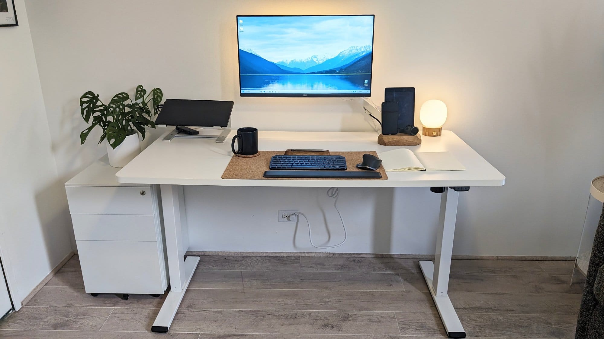 A home office setup featuring a standing desk with a dual-monitor arrangement, a keyboard and mouse on a cork mat, a mug, a notebook, a spherical lamp, and a monstera plant