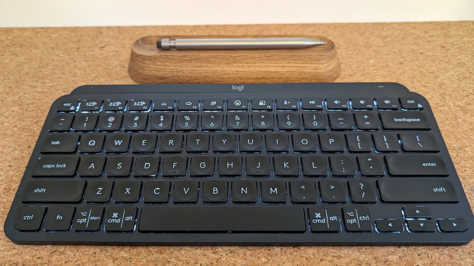  A close-up image of a black Logitech keyboard with illuminated keys, placed on a cork surface with a wooden pen holder behind it