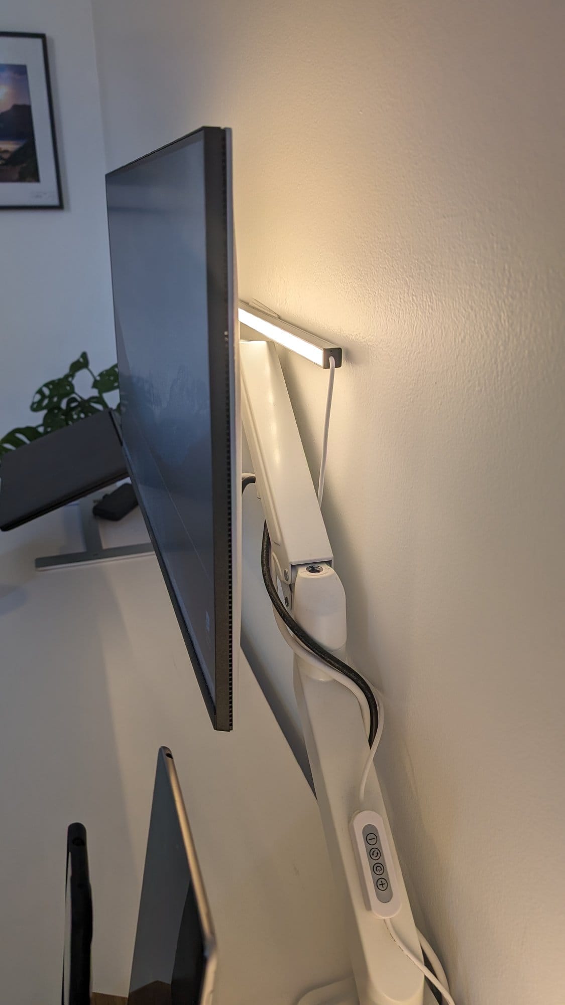 A side view of a thin monitor attached to a white articulated arm mount, with a power strip button visible, against a white wall, and a framed picture in the background