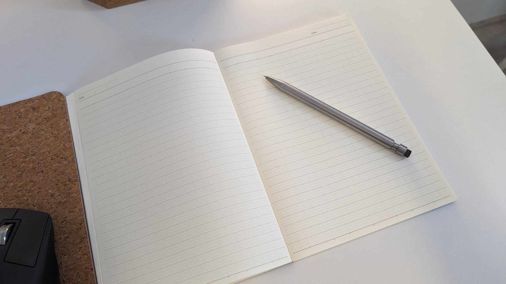  An open lined notebook with a metallic pen resting on the right page, placed on a white surface next to a cork desk mat