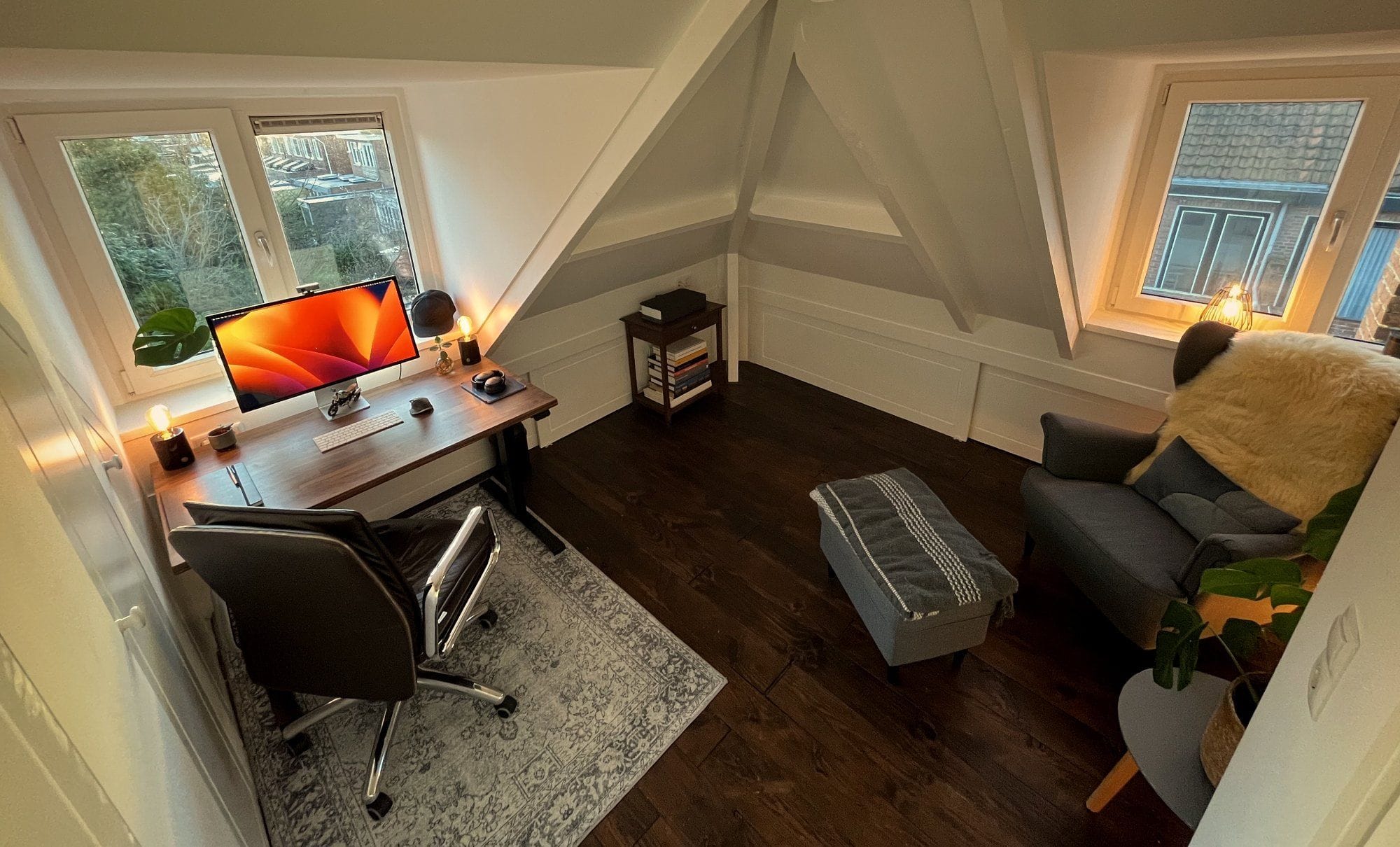 A tranquil attic home office setup with a dark wood floor, a traditional rug under a contemporary desk and chair, ambient lighting, and a cosy armchair with a fur throw