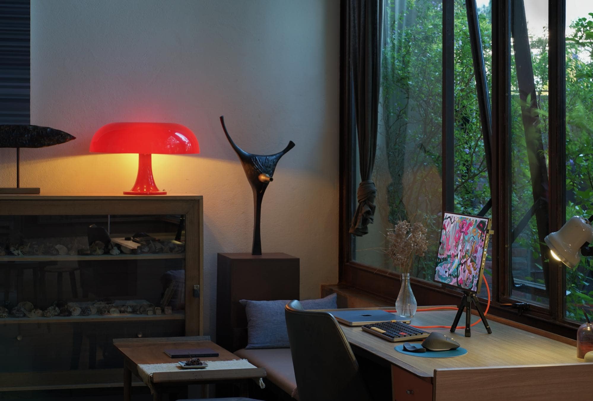 An intimate desk space bathed in the warm glow of a red table lamp, adjacent to a window with a view of lush foliage