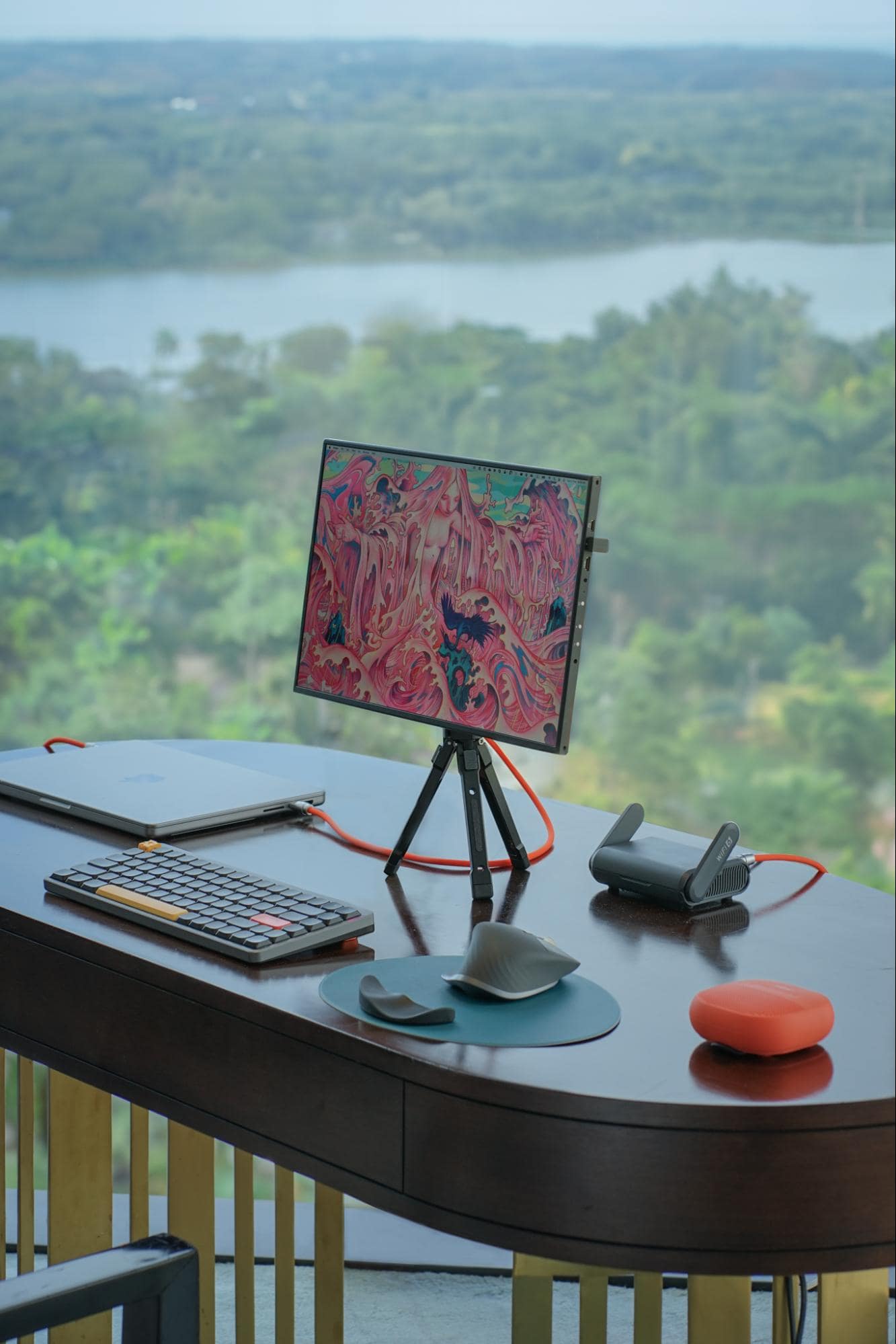A serene desk setup with a scenic backdrop, featuring a laptop, a graphic tablet on a tripod stand, a mechanical keyboard, and a portable speaker, all neatly arranged on an oval table with a picturesque lake and lush greenery in the distance