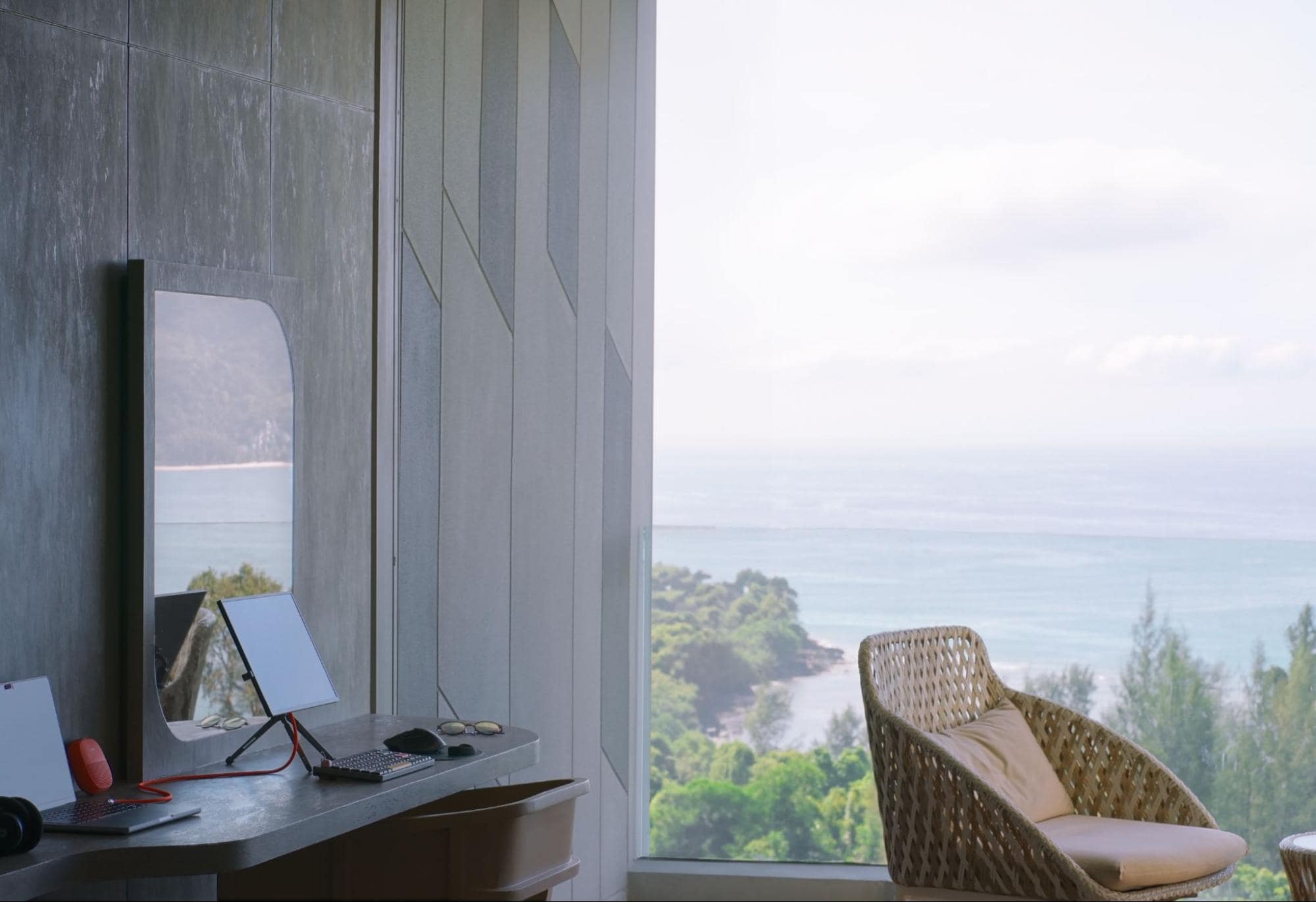 An airy desk setup with a coastal view, featuring a tablet on a stand, a mechanical keyboard, and personal items, complemented by a comfortable wicker chair