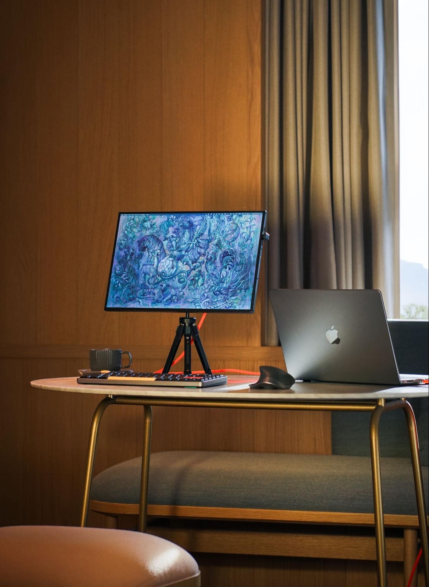 An elegant and modern desk setup featuring a MacBook, a graphic tablet propped on a tripod stand, a mechanical keyboard, and a coffee cup, all placed on a stylish oval table against a warm wooden backdrop