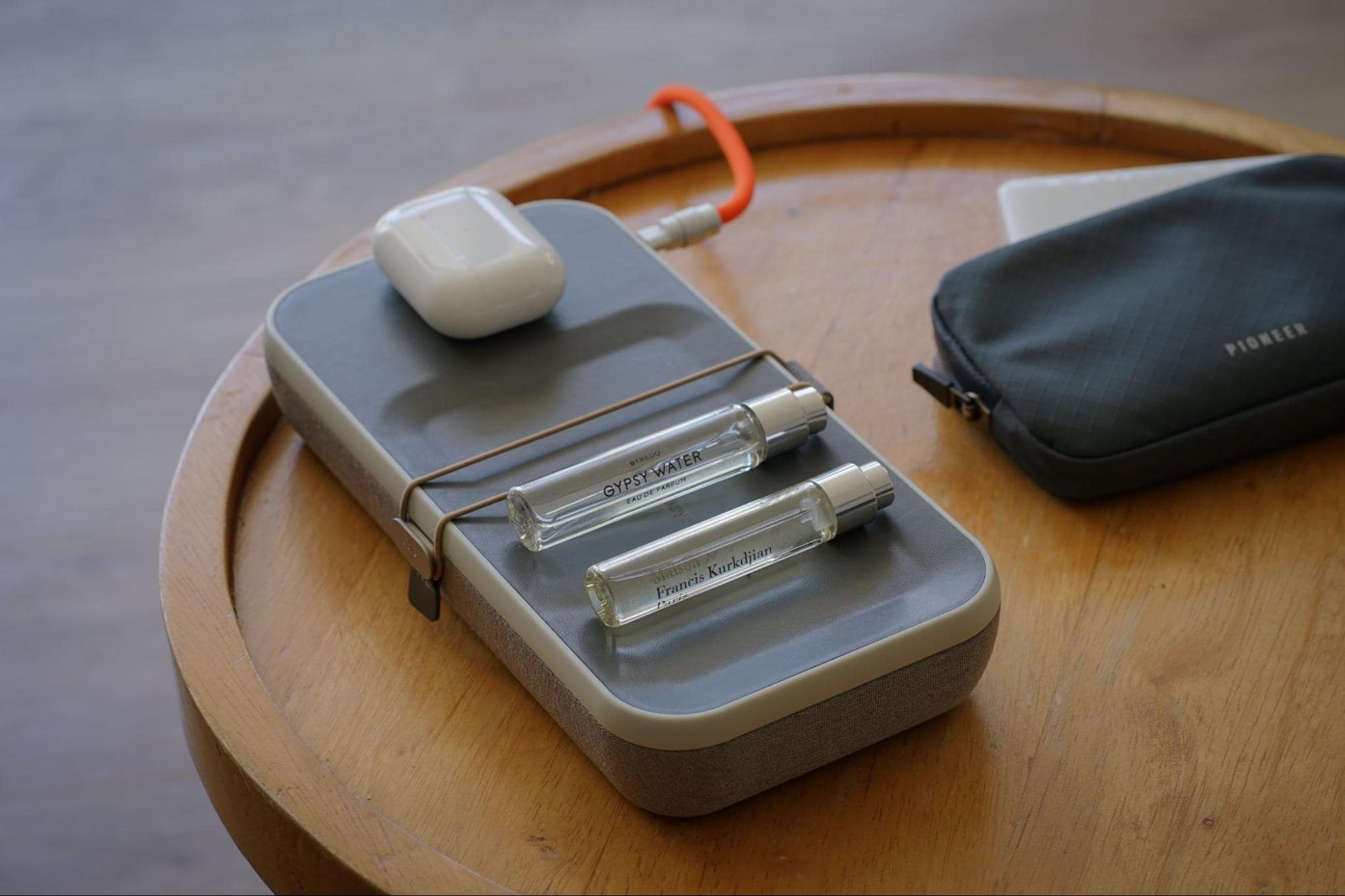 A close-up of a neatly arranged selection of personal items on a wooden table, including a pair of wireless earbuds, two sleek perfume vials, and a portable tech accessory pouch