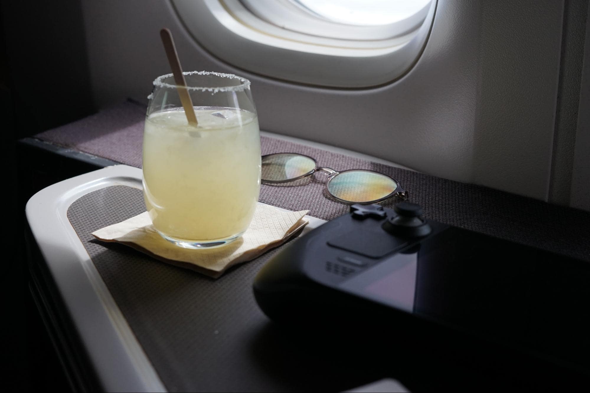A refreshing beverage on an airplane tray table beside a pair of sunglasses and a handheld gaming device Steam Deck, capturing a moment of relaxation during a flight
