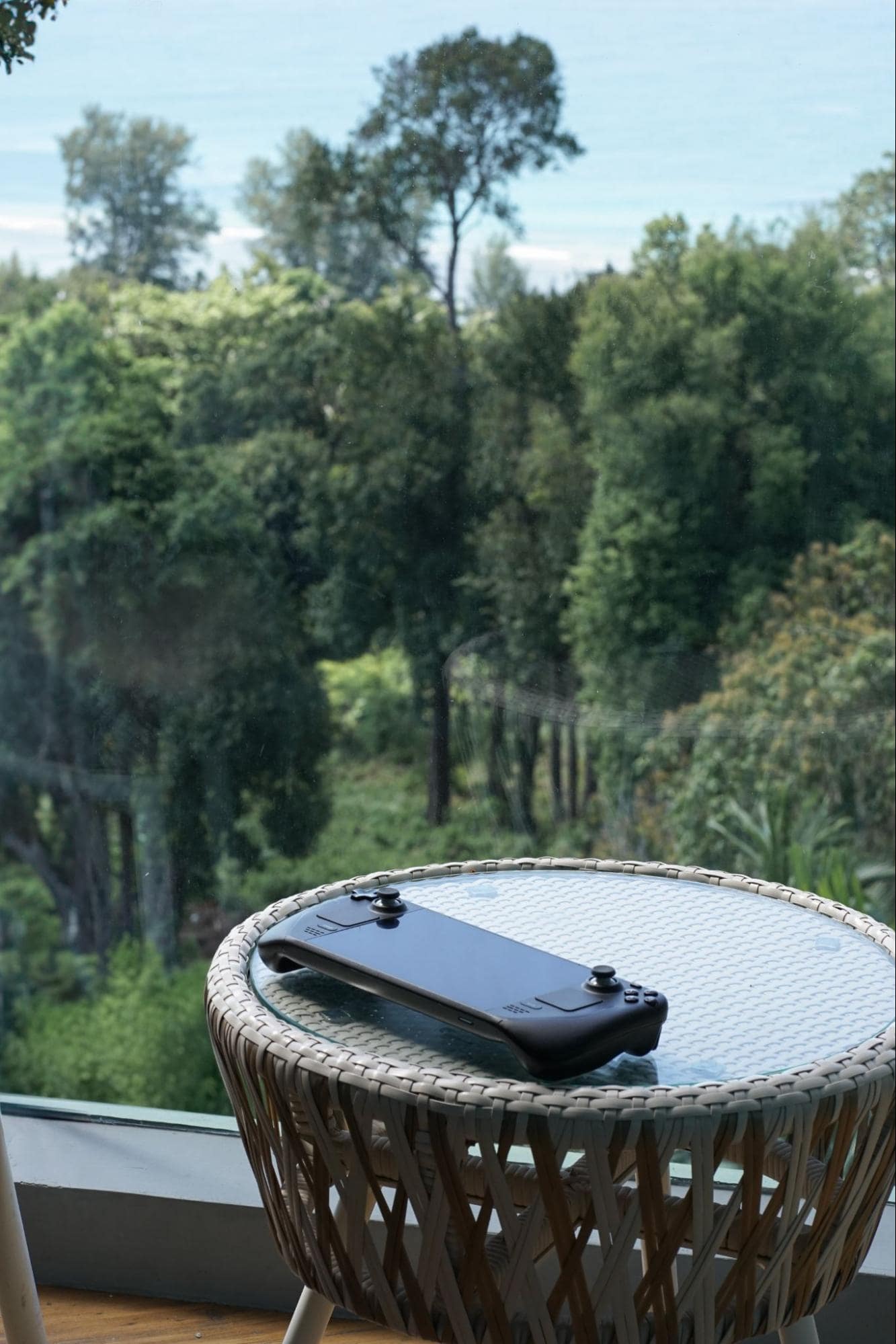 A handheld gaming console Steam Deck resting on a round wicker side table, with a serene view of the ocean and lush greenery through a large window, offering a peaceful spot for entertainment and relaxation