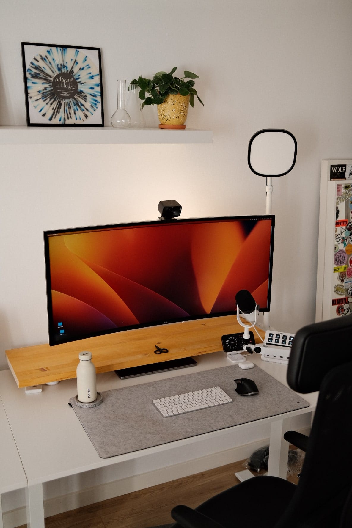 A modern workspace featuring a large monitor with a webcam on top, a ring light, a desk mat with keyboard and mouse, and a shelf with art and a potted plant above