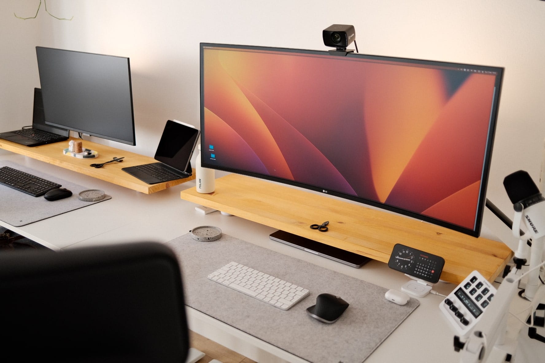 A home office setup with two monitors on a wooden stand, a laptop, a webcam, and a microphone, with a keyboard and mouse on a grey desk mat