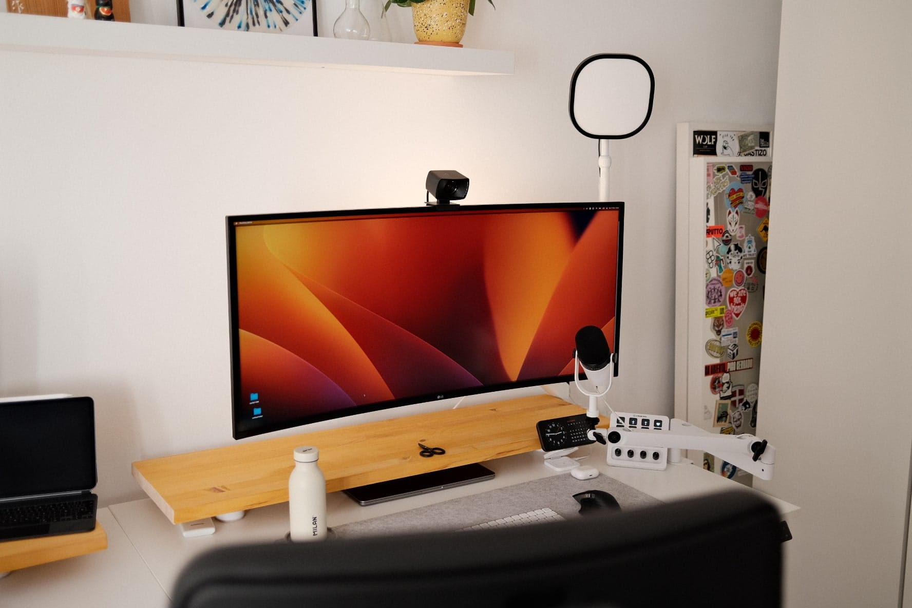 A desktop monitor with a webcam on top, a microphone on an adjustable arm, a ring light, and various desk accessories against a wall with decorative items