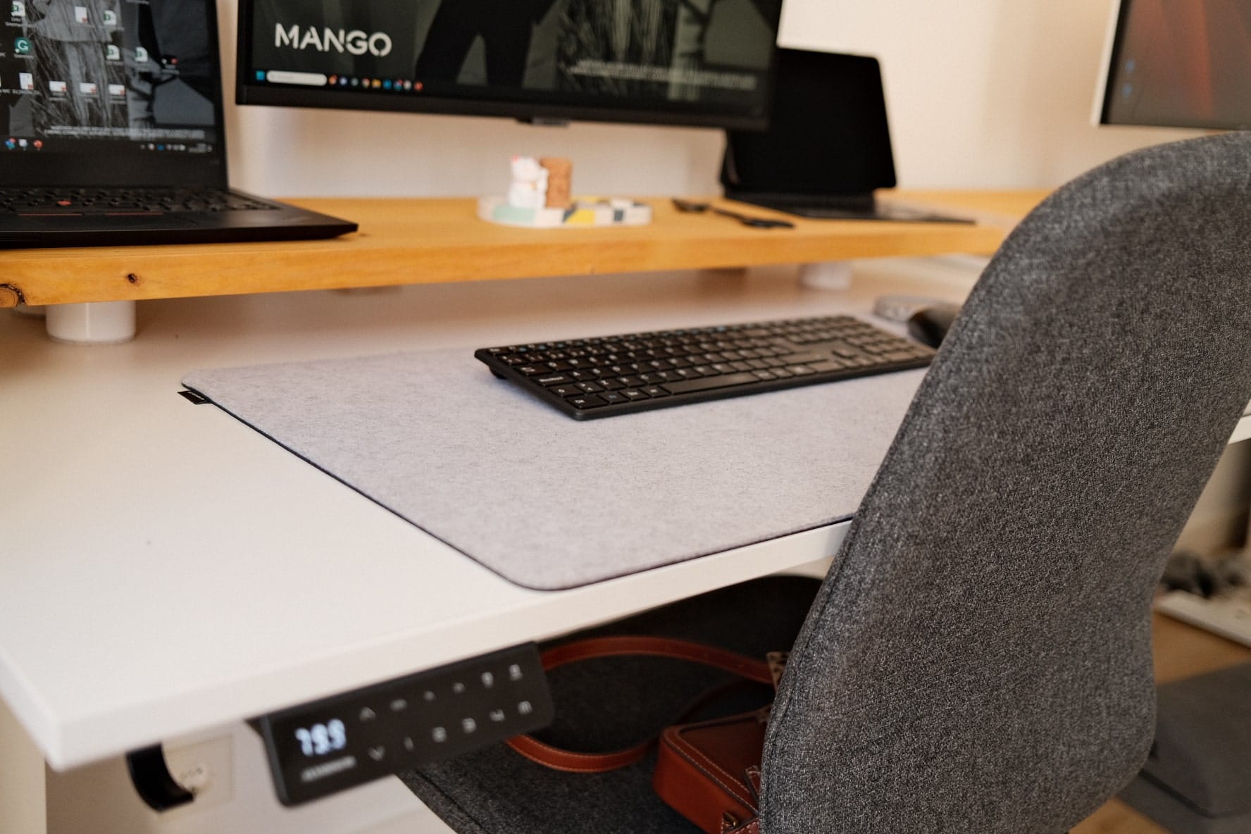 A home office setup with a keyboard on a felt mat, a wooden monitor riser with a laptop on top, and a grey upholstered office chair