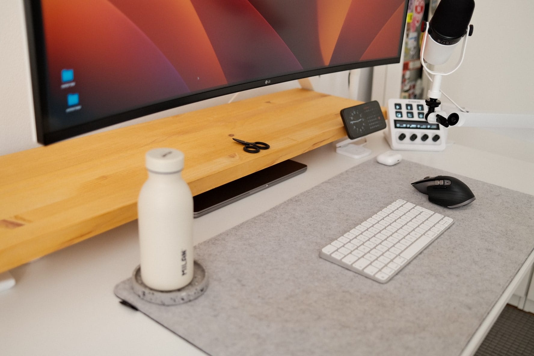 A tidy desk featuring a large monitor, white keyboard, ergonomic mouse on a grey mat, wooden monitor stand with a laptop underneath, microphone on a boom arm, and a reusable water bottle