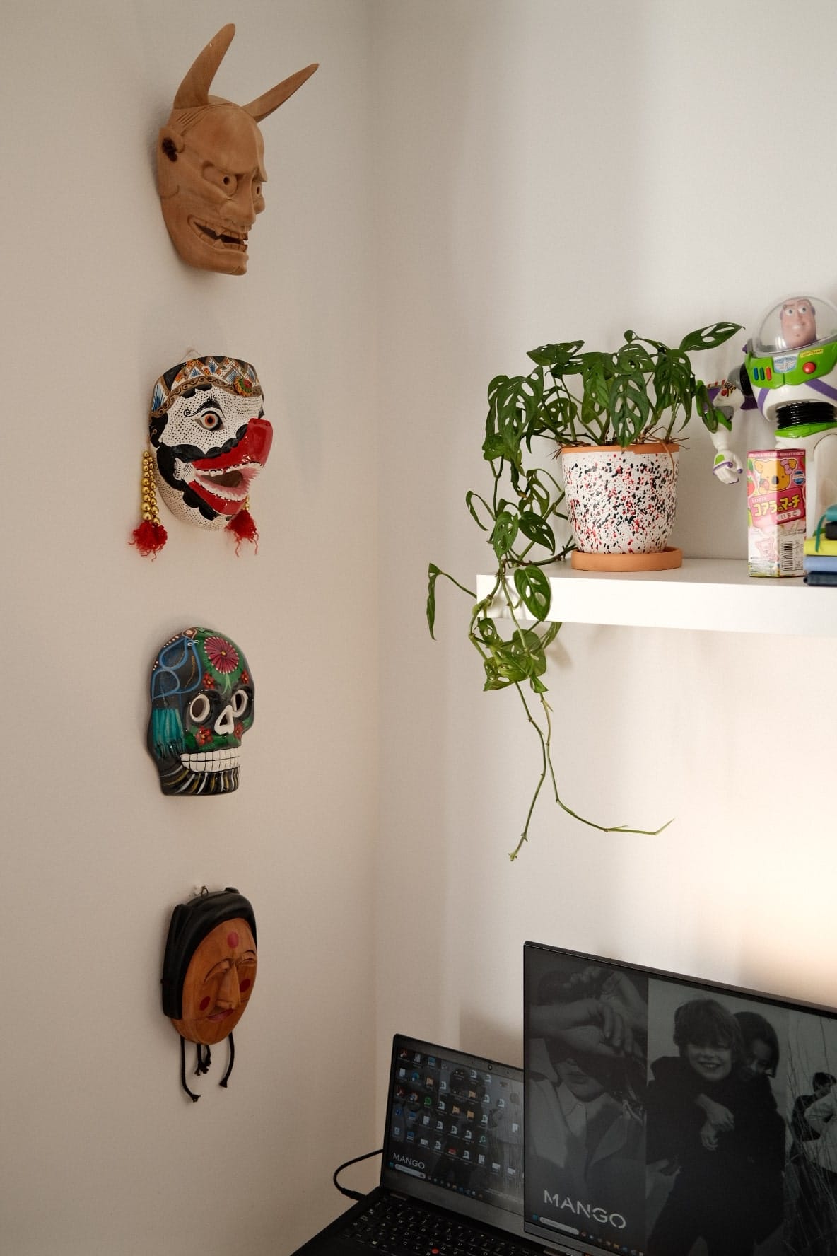 Part of a room with cultural masks hanging on the wall, a potted plant on a shelf, and a laptop with a screensaver on a desk