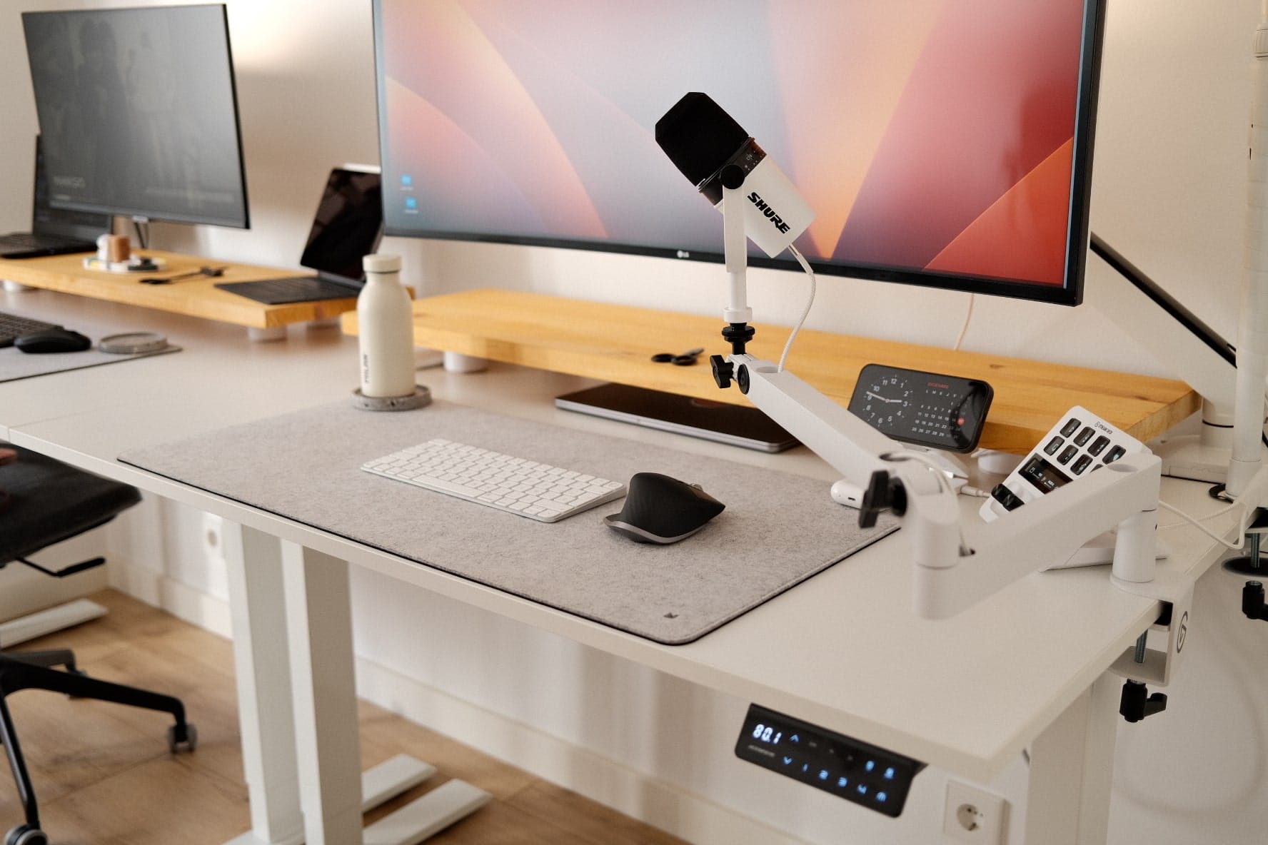 An organised desk space with a dual monitor setup, microphone on a white boom arm, white keyboard, ergonomic mouse on a felt desk mat, and a streaming control deck