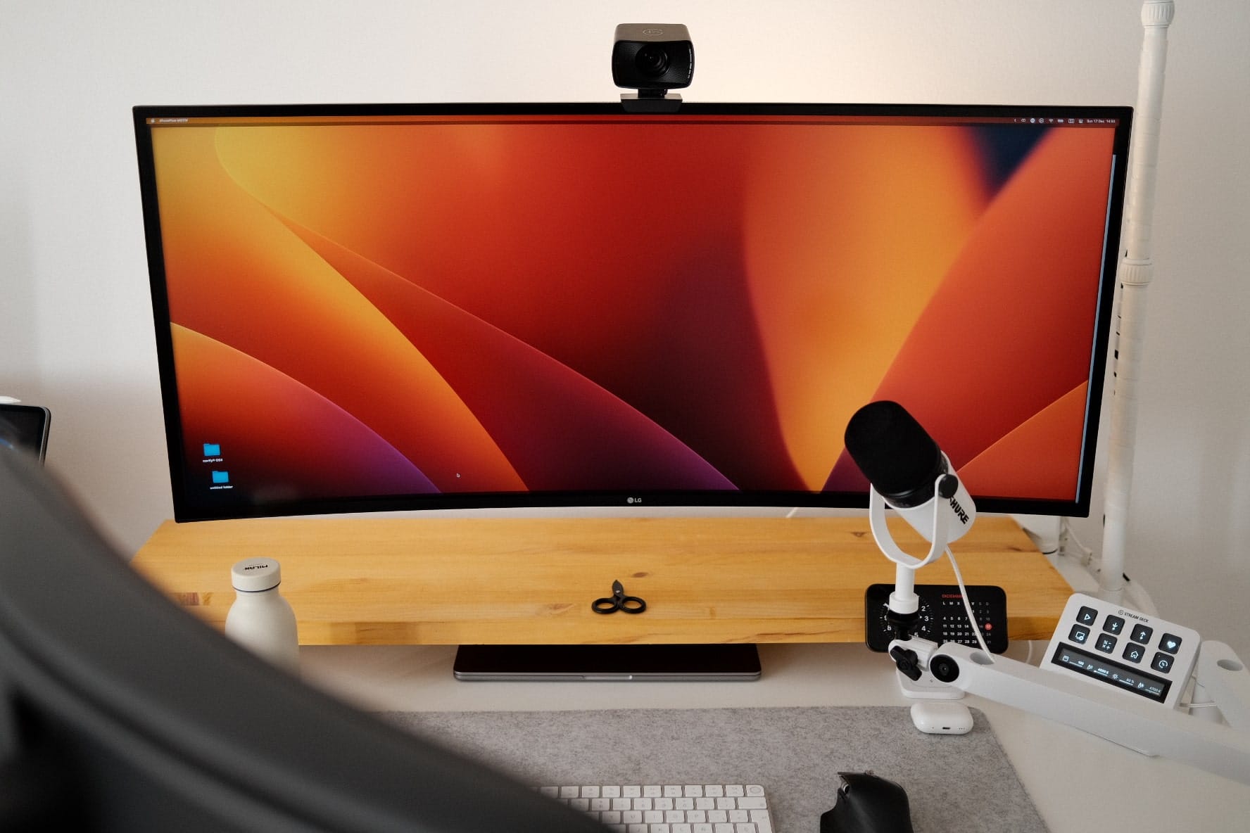 A computer monitor displaying a vibrant wallpaper with a webcam on top, a microphone to the side, and a wooden stand with scissors on it, on a white desk