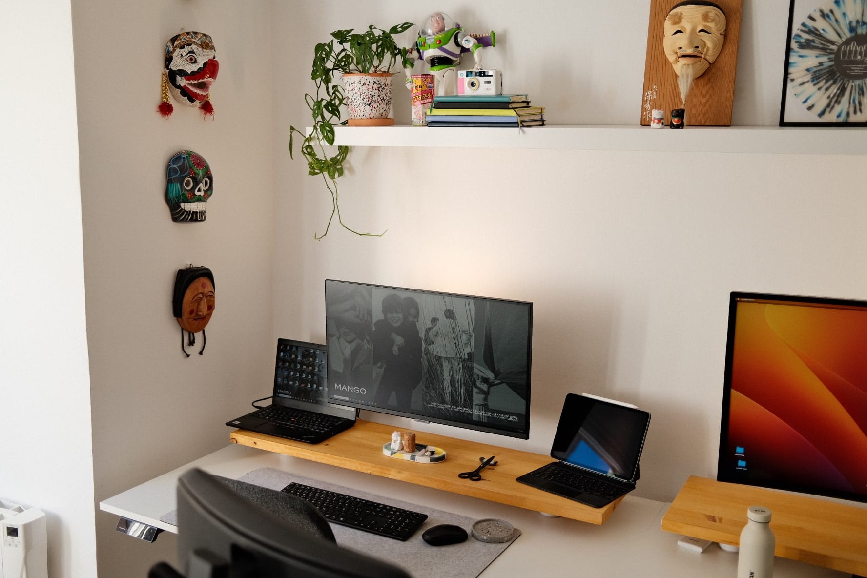 A home office setup with a dual monitor display, decorative masks on the wall, a shelf with a plant and toys, and a desk with various work items