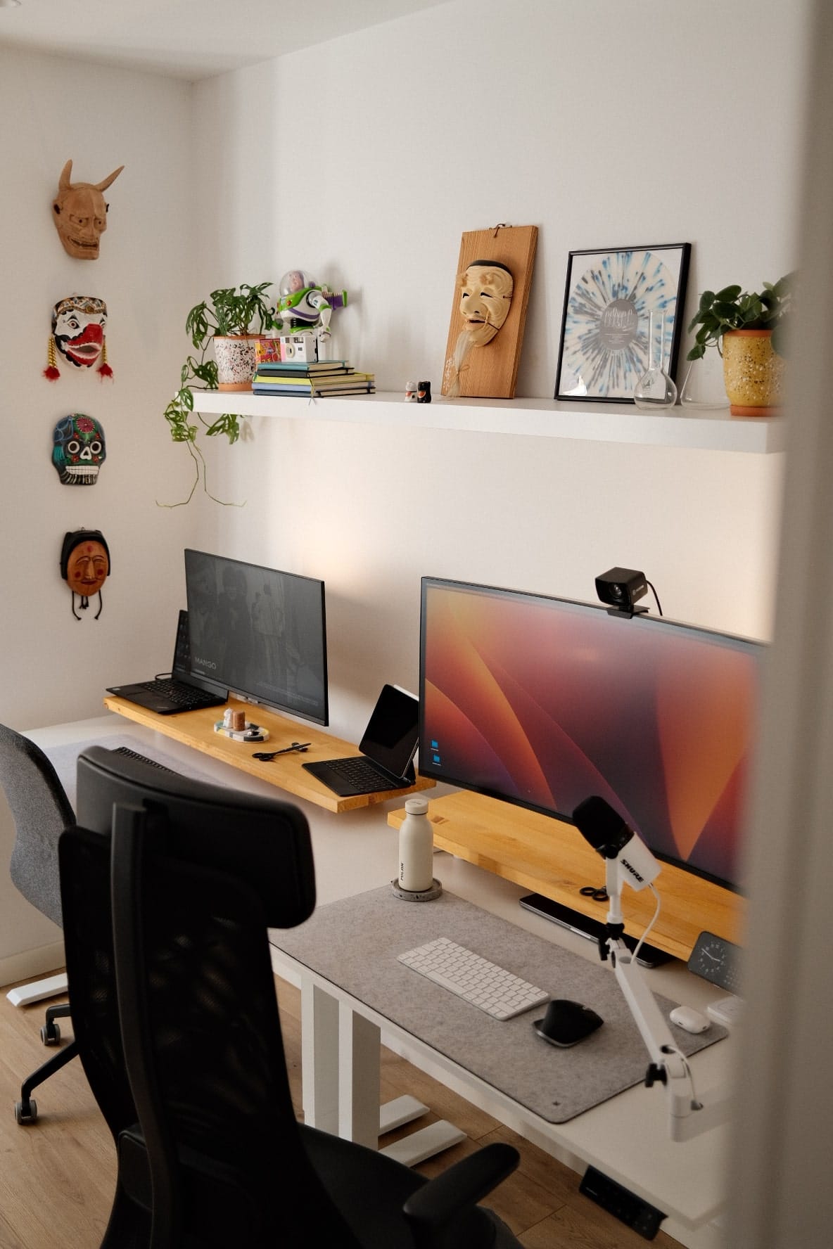 A cosy shared home office setup with two ergonomic chairs, two monitors, a laptop, microphone, and decorative masks and plants on the shelf